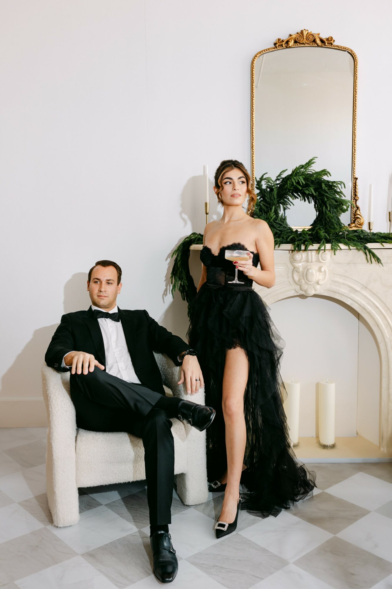A woman in a black gown and a man in a tuxedo sit and stand near a fireplace with a mirror and greenery, in a decorated room.