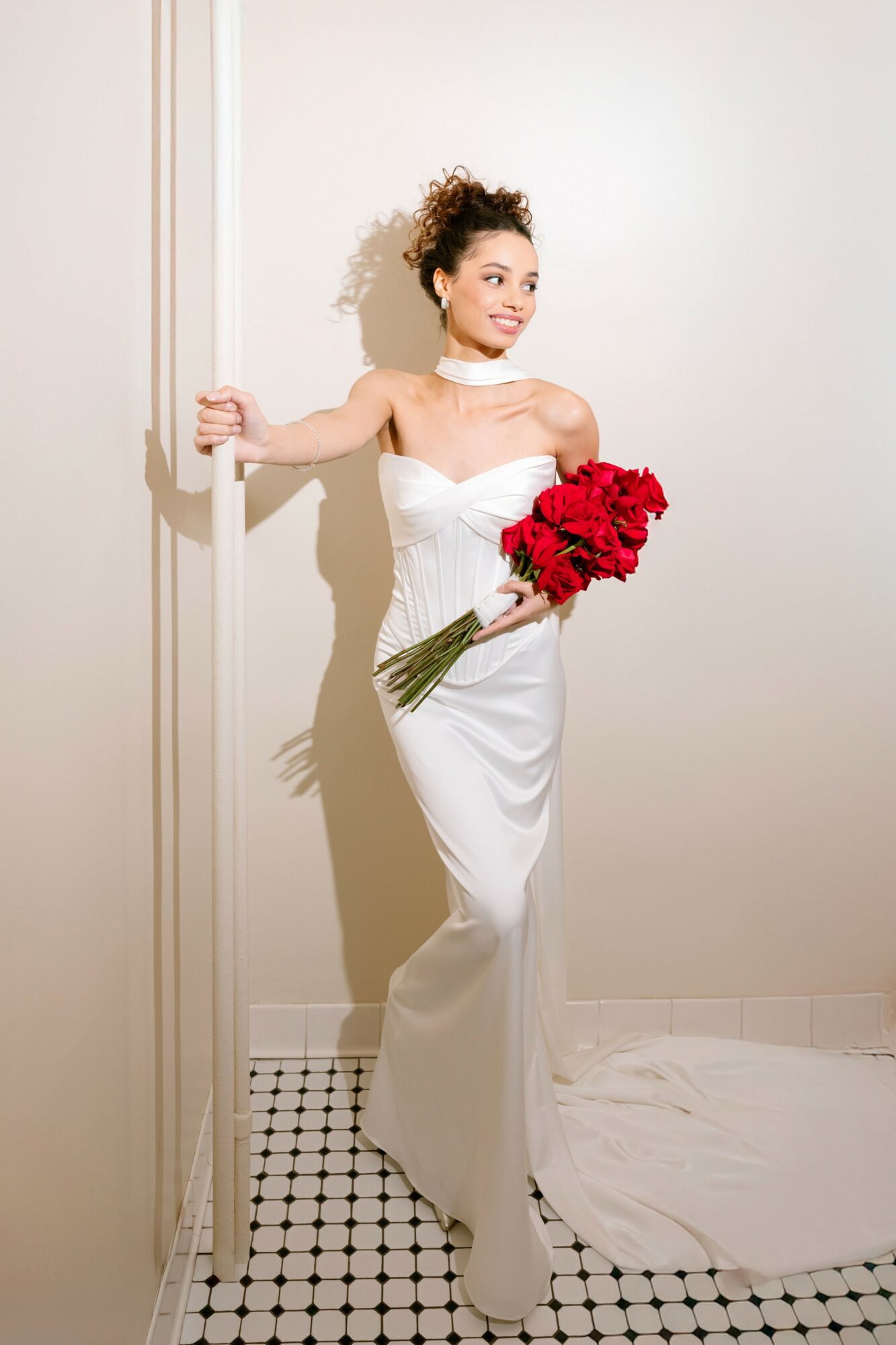 Woman in white dress holding a bouquet of red flowers, standing against a white wall, smiling, with a train on the floor.