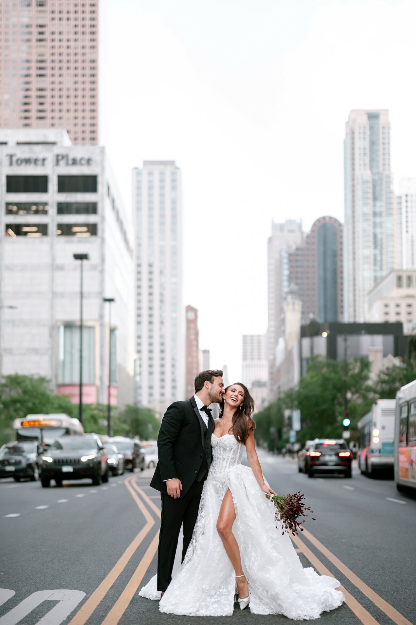 A couple in wedding attire standing in the middle of a city street with tall buildings and cars around them.