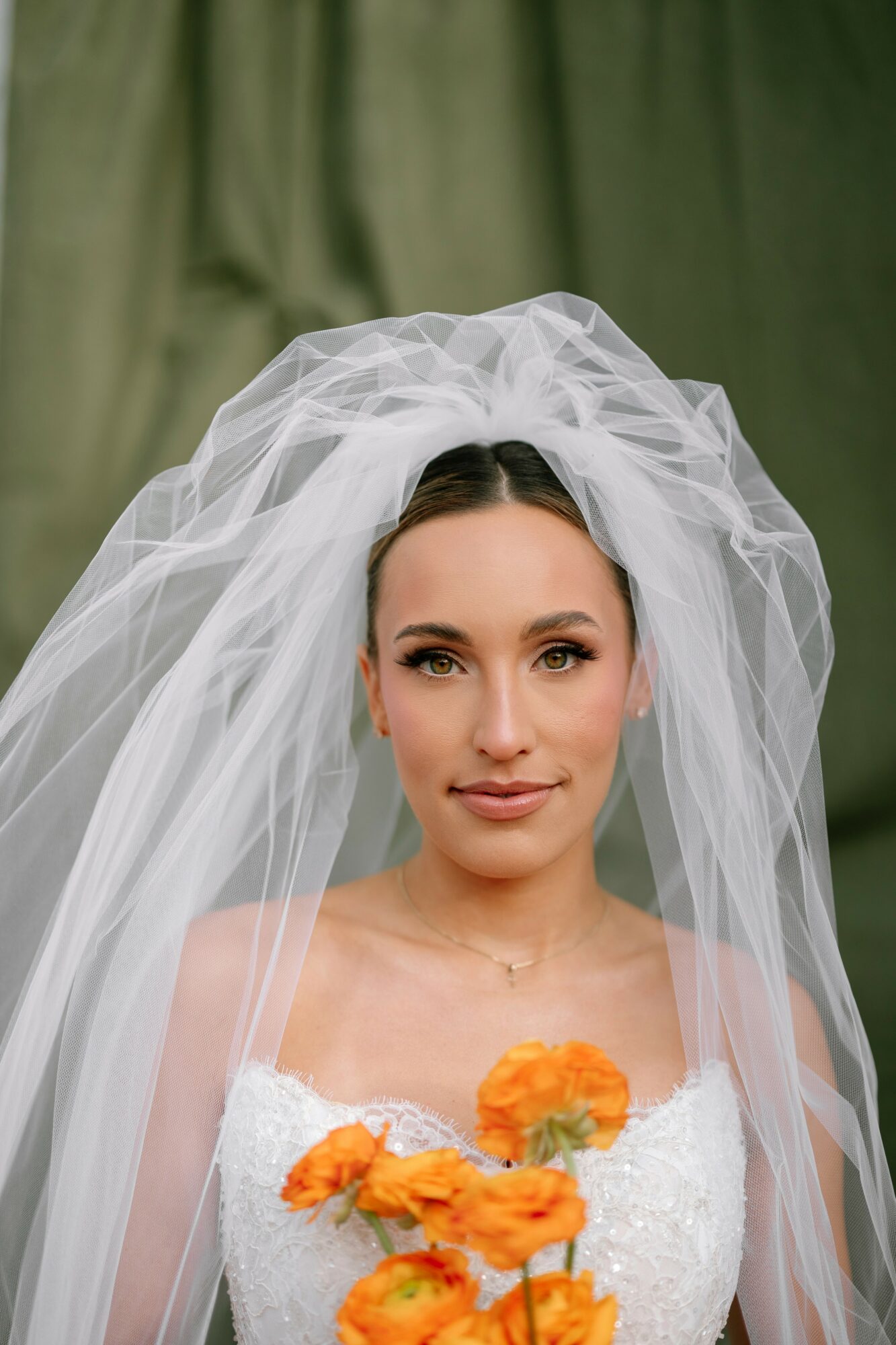 Bride with veil and white dress holding orange flowers, looking at camera, green background