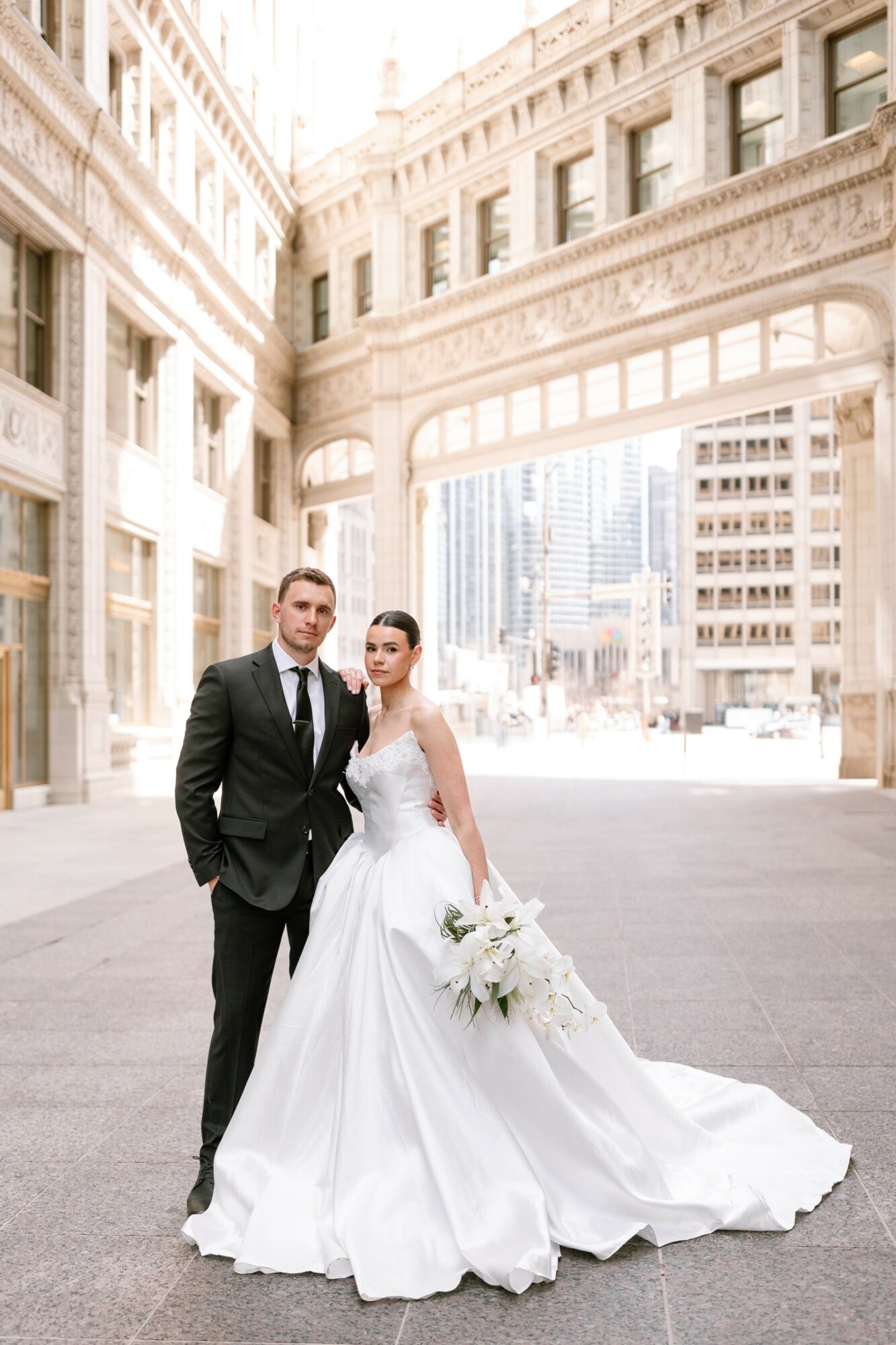 Bride and groom in wedding attire standing outdoors in an urban setting with tall buildings and an archway behind them.