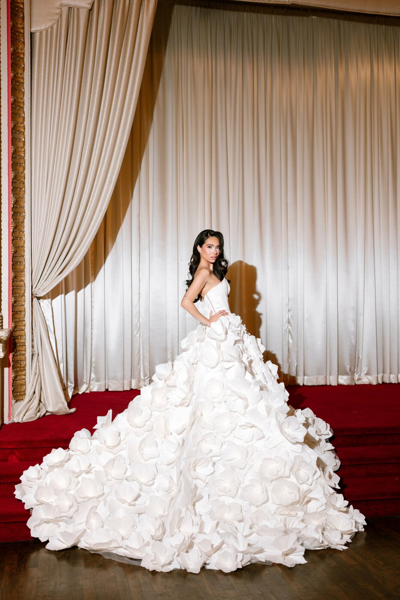 Woman in wedding dress standing on stage with large curtain backdrop, red carpet, and wooden floor.