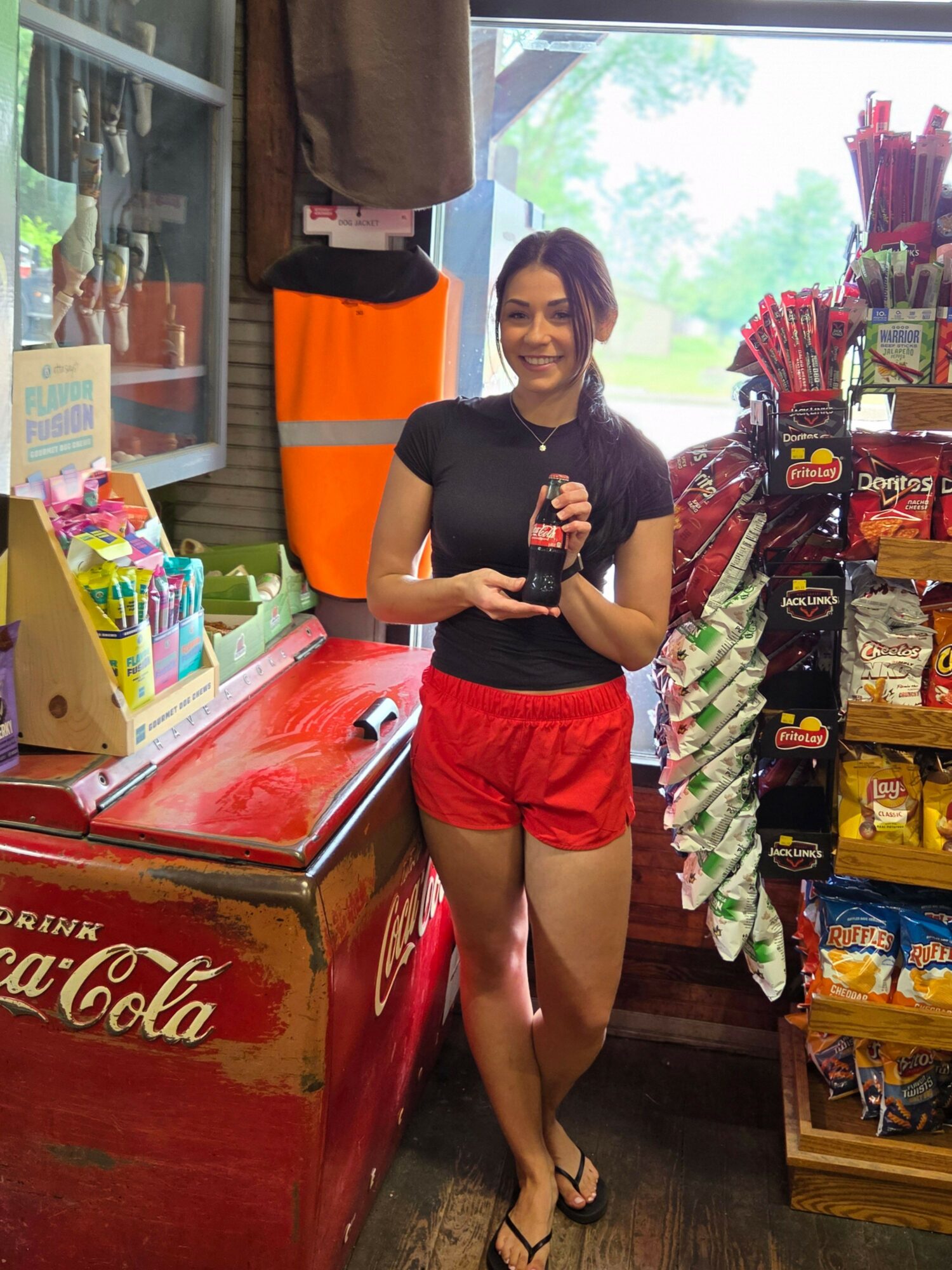 Young woman with dark hair in a black shirt and red shorts standing inside a store, holding a beverage, surrounded by snacks and drinks.