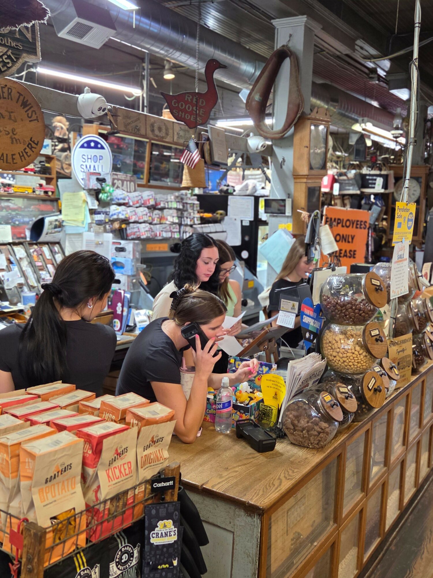 Three women work behind a wooden counter in a store, surrounded by jars, boxes, and decorative items.