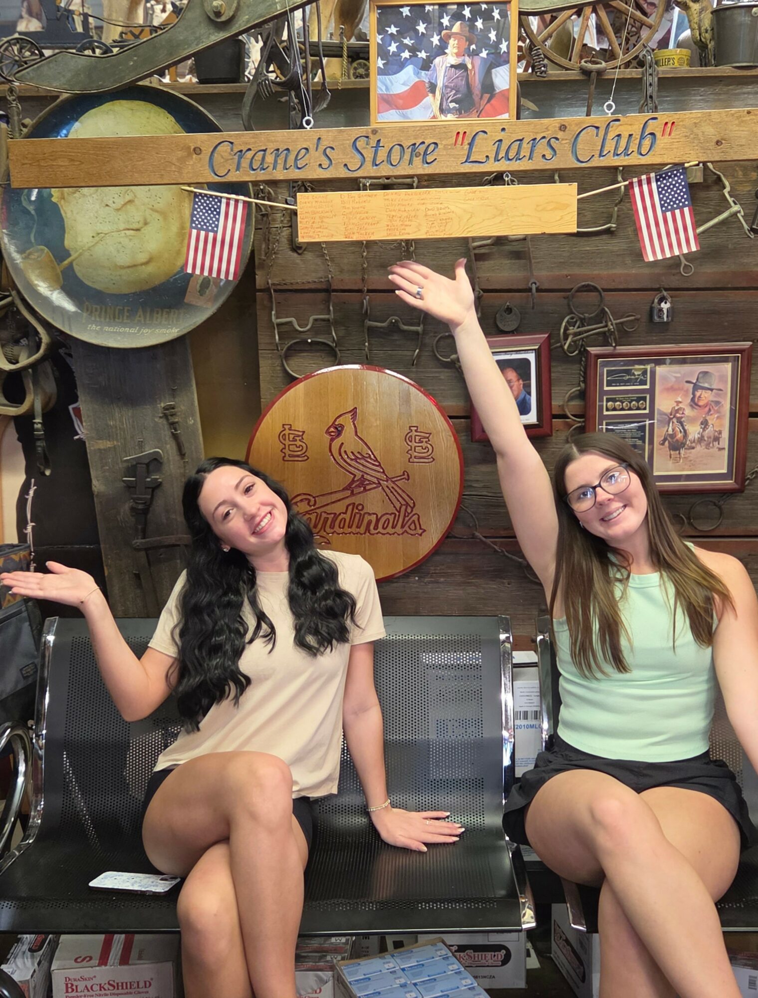 Two women sitting on a bench, smiling, with one raising her arm, in a decorated room with American flags and signs.
