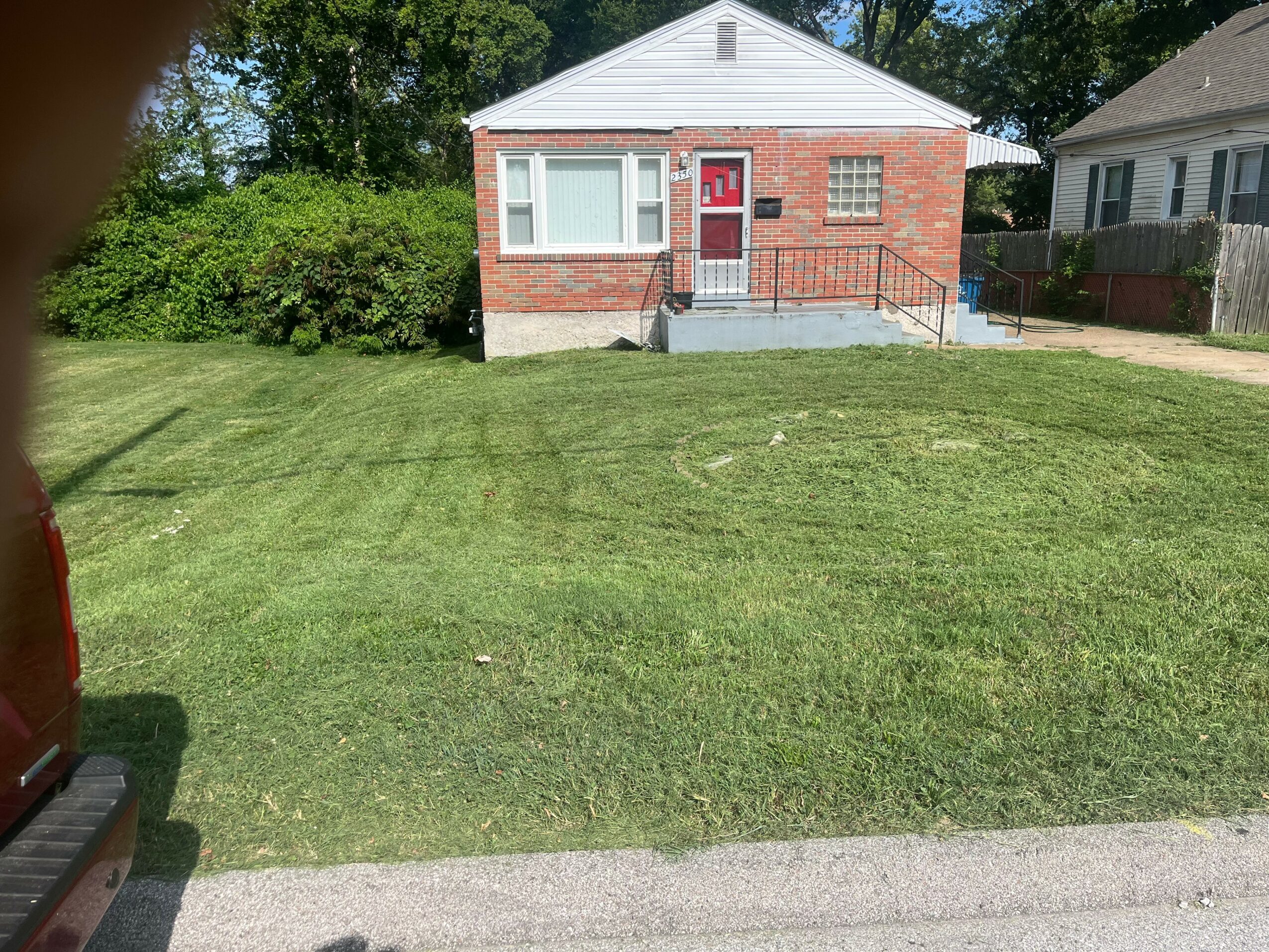 Front view of a small brick house with a white upper section, red door, and steps, surrounded by a green lawn.