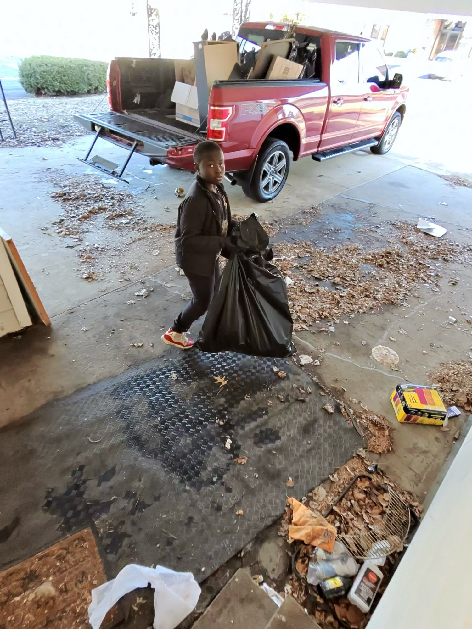 Child holding a large black trash bag standing on a dirty floor with scattered debris, a red pickup truck loaded with boxes in the background.