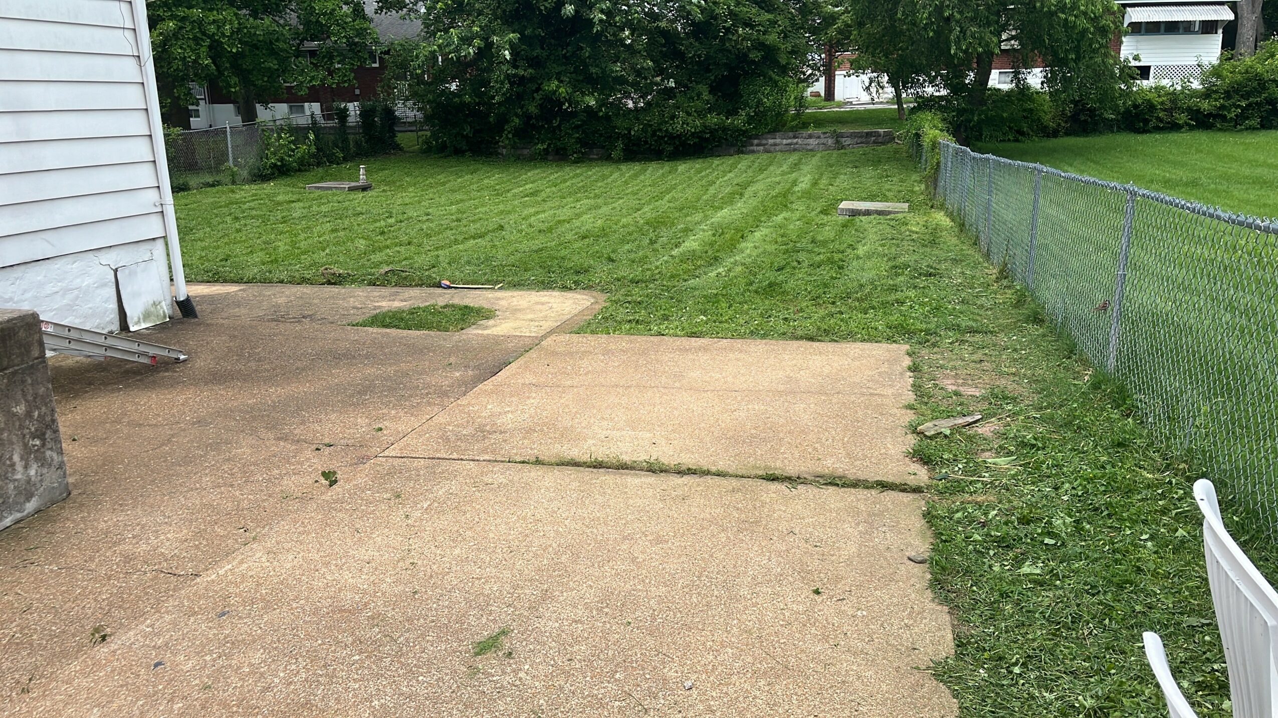 Concrete patio with grass and a chain-link fence in the background, next to a white building, with a white chair partially visible.