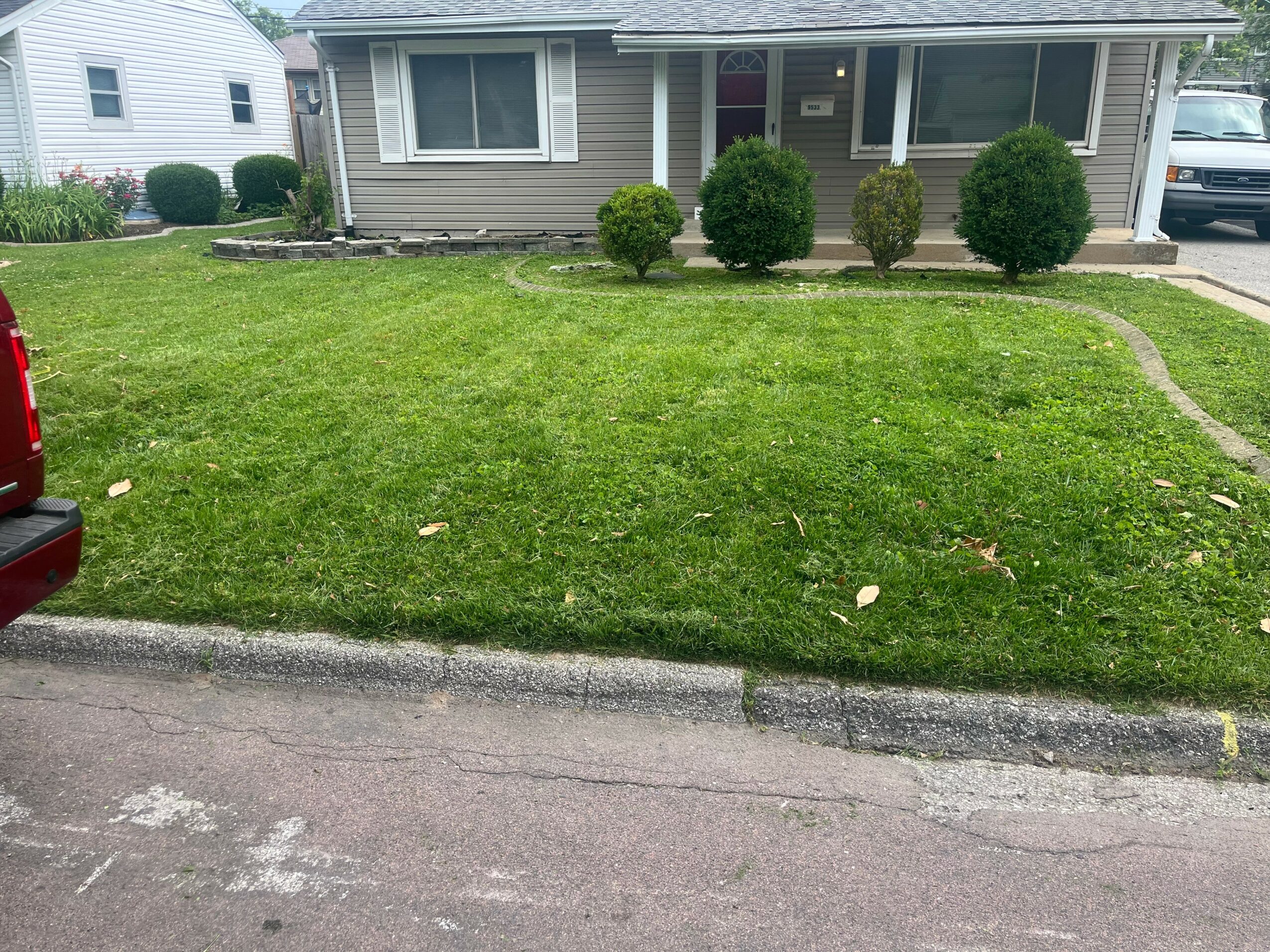 Front yard with green lawn, four bushes, house with porch, driveway, and parked car, viewed from street.