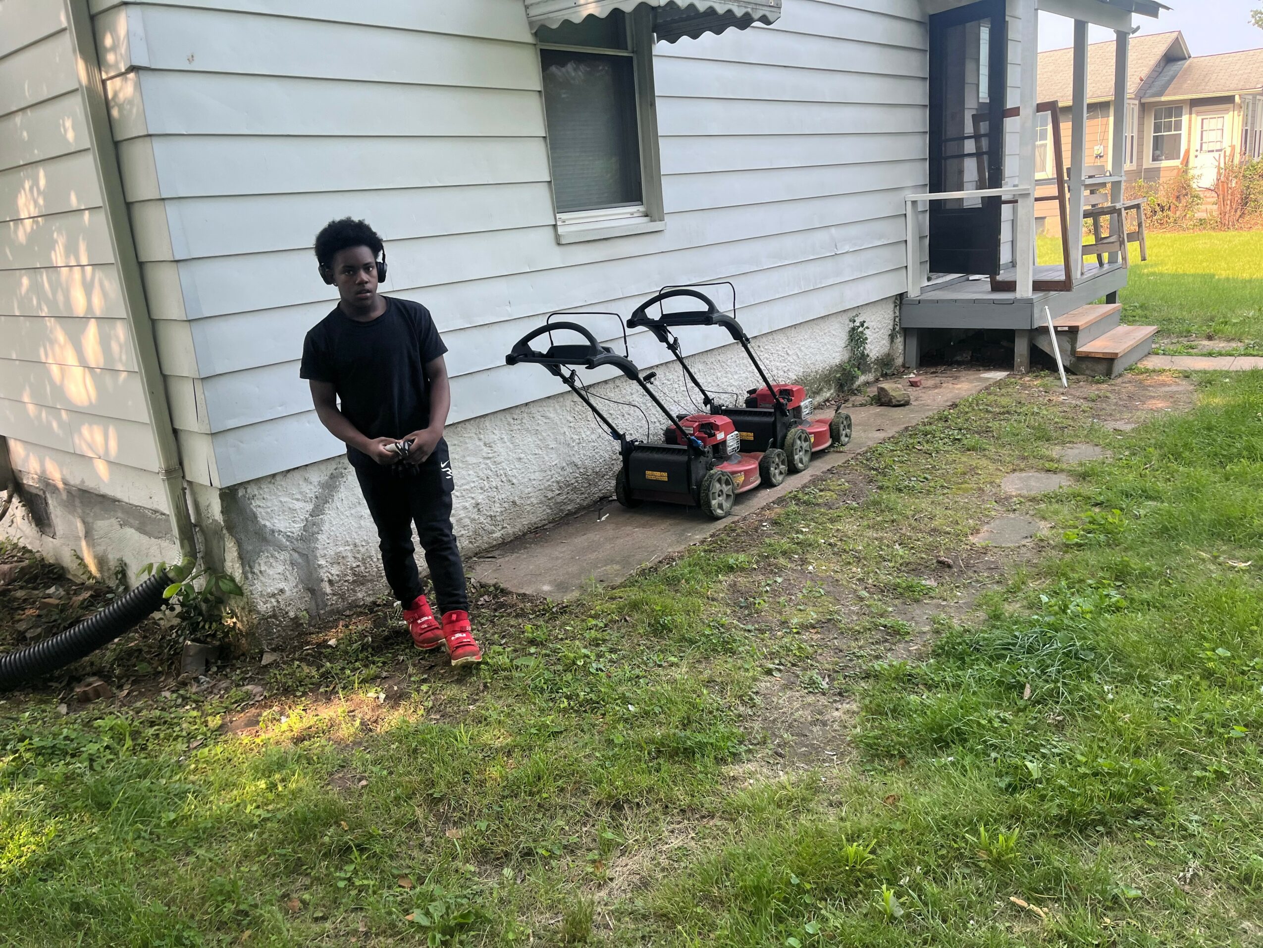 A boy stands outside a house with two lawnmowers on the ground nearby, grass and a porch in the background.
