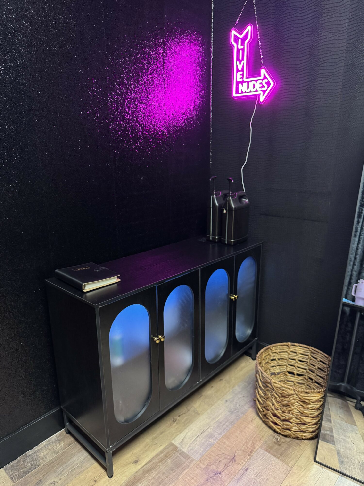 Black cabinet with glass doors and blue lighting, a woven basket on the floor, and a neon sign on the wall.
