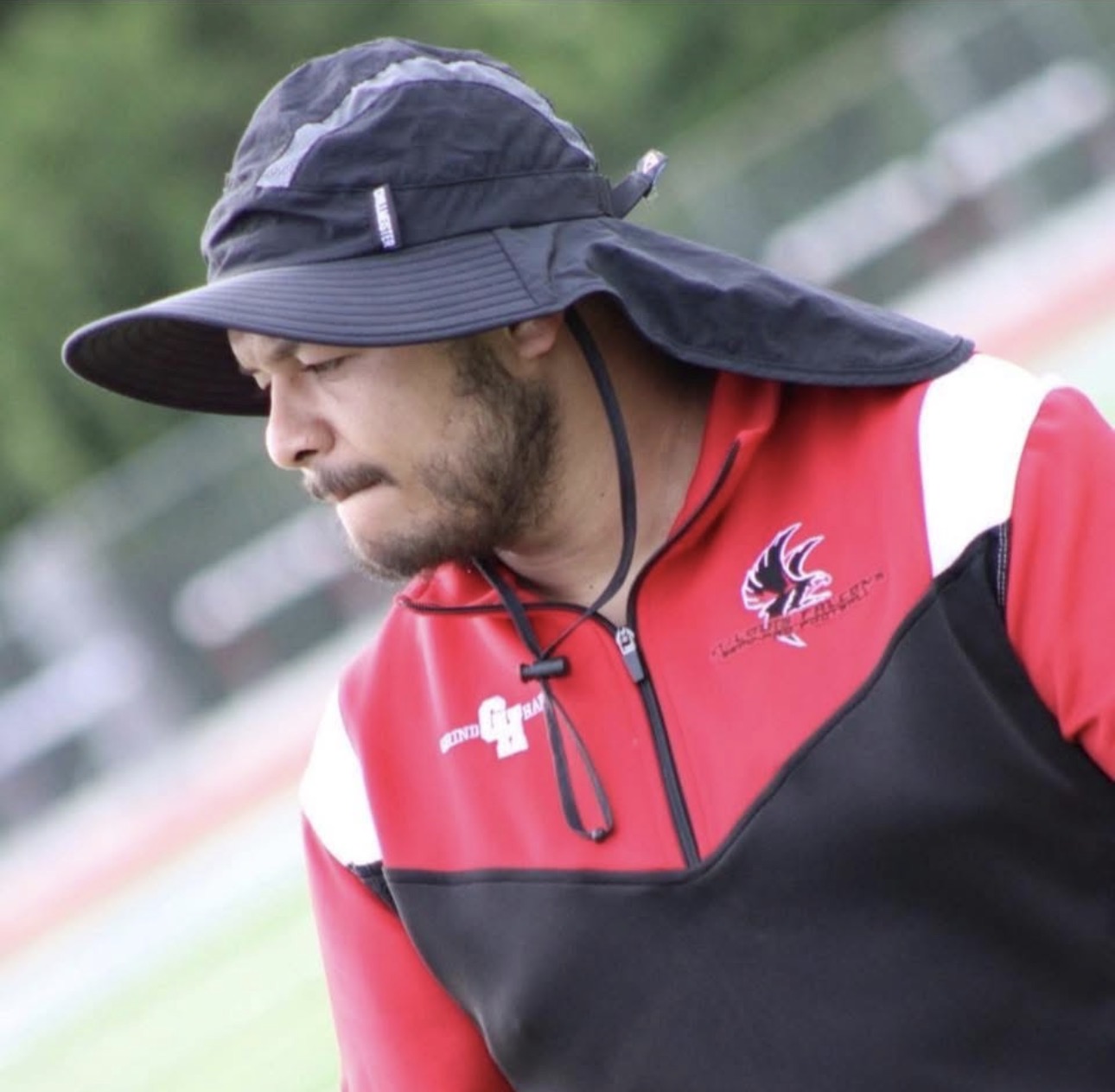 Man wearing a wide-brimmed hat and red sports jacket outdoors, looking down, with a blurred background.