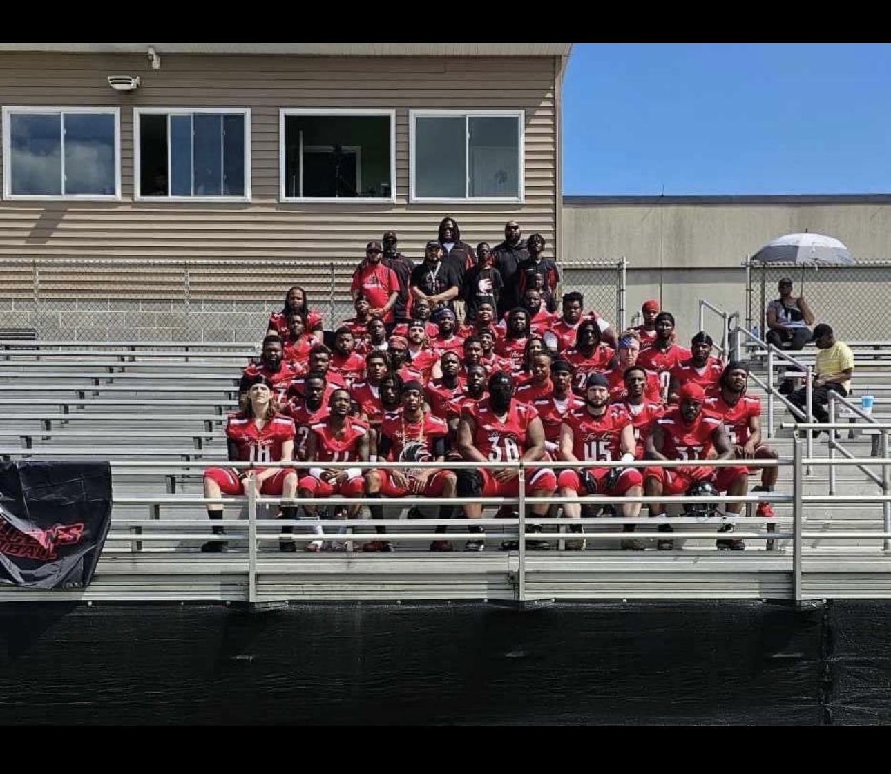 Group of football players and coaches sitting on bleachers outdoors, wearing red uniforms, with a building and blue sky in background.