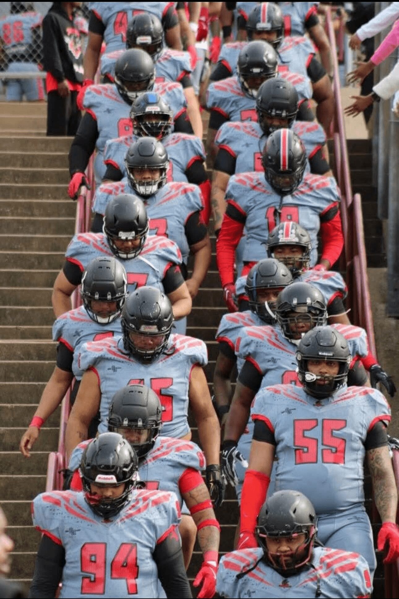 Football team in gray uniforms with red accents and black helmets walking down stairs, some with numbers visible.