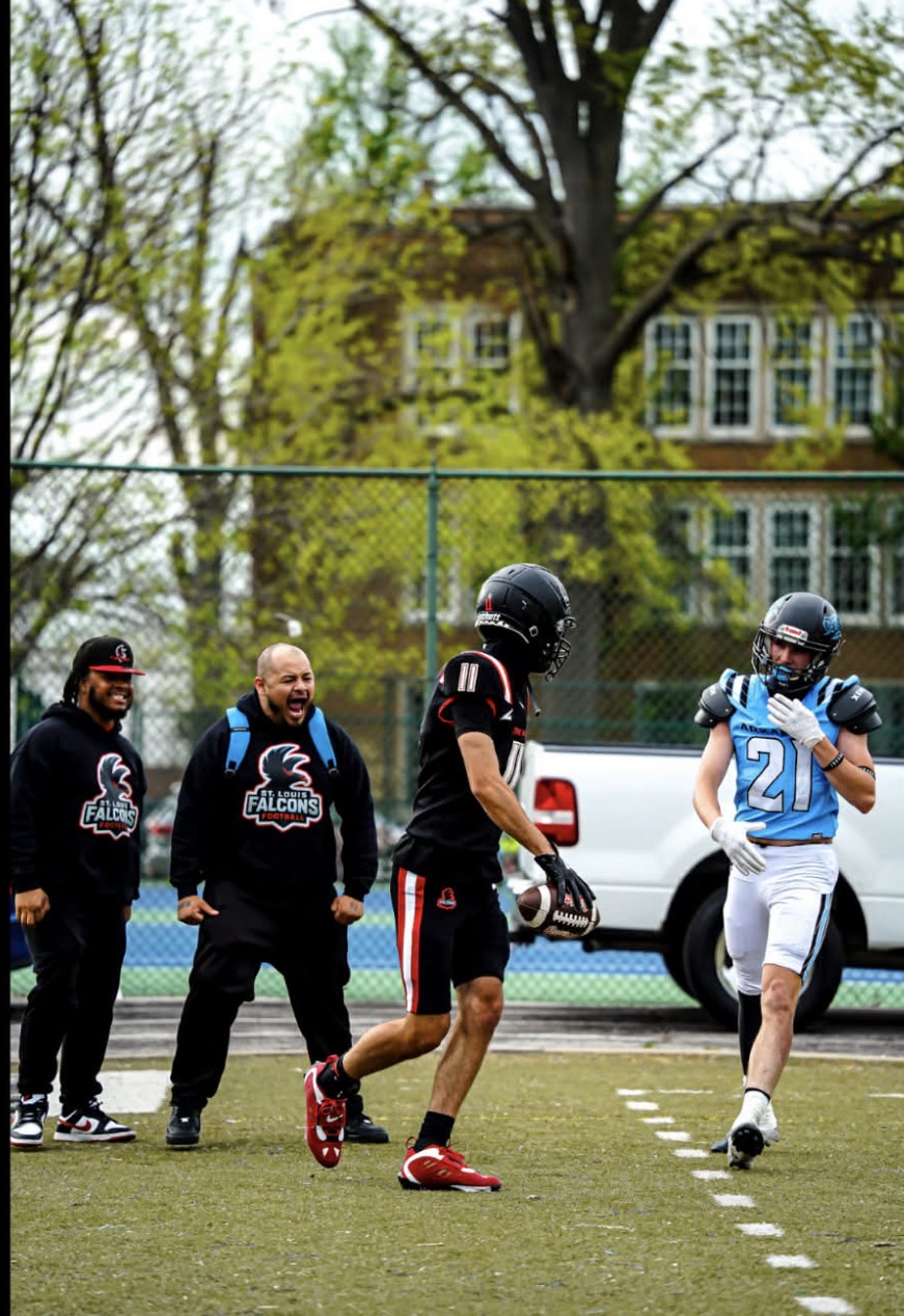 Four people on a football field, one in a football uniform, three in casual clothing, with trees and a building in the background.