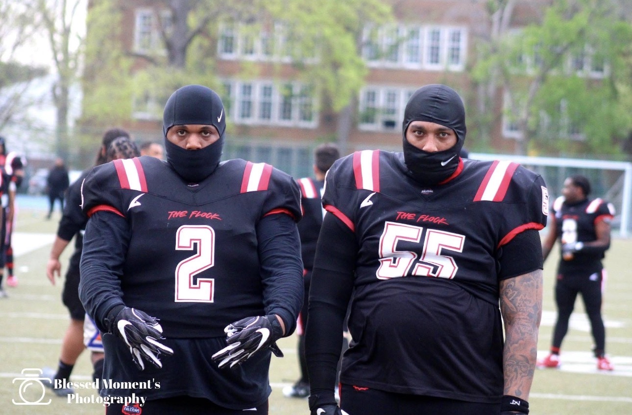 Two football players in black uniforms and masks stand outdoors with trees and buildings in background.