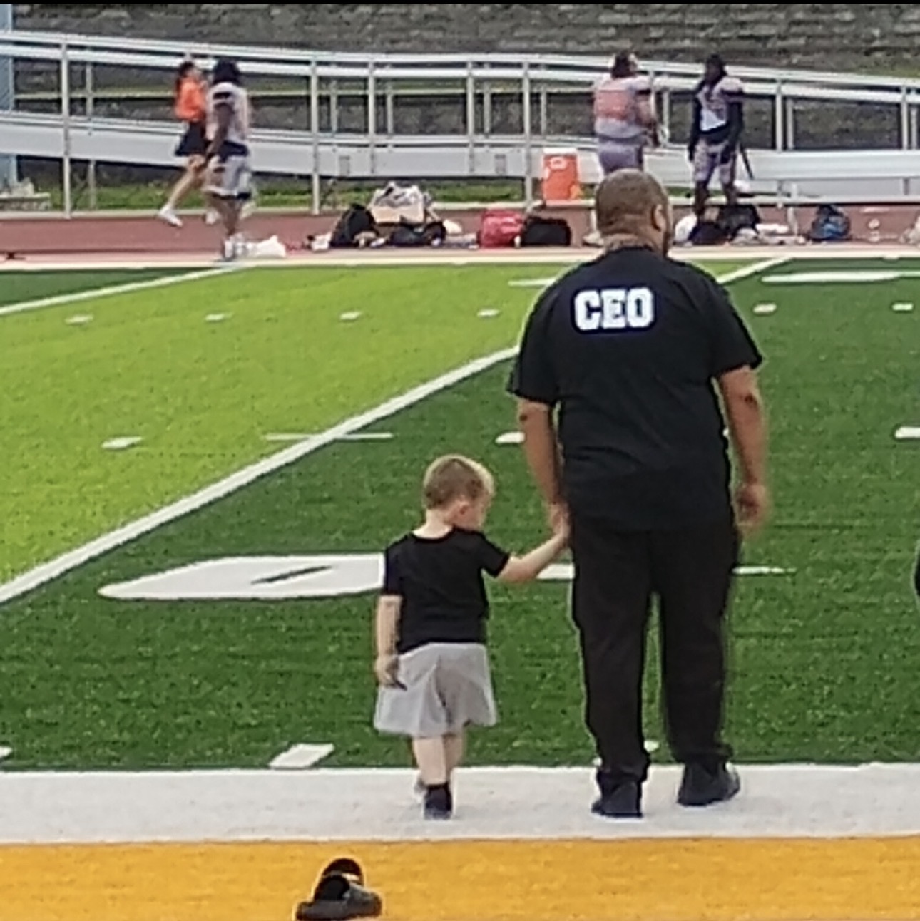 Child holding hands with a man wearing a 'CEO' shirt on a sports field, with people in the background.