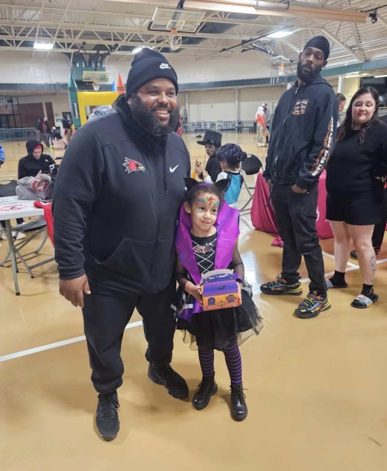A man and a young girl stand together in a gymnasium, smiling, with other people in the background.