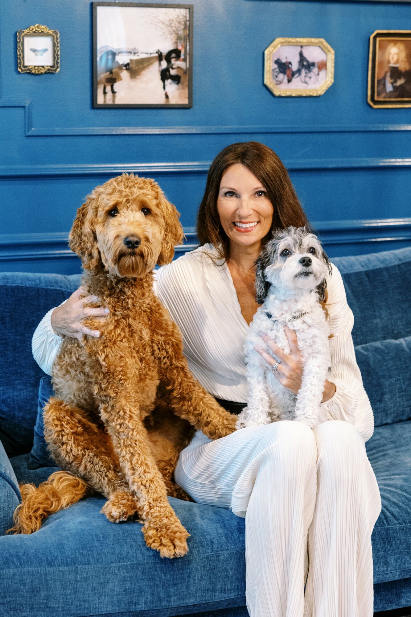 Woman smiling with two dogs on a blue couch in front of framed artwork, woman wearing white striped outfit.