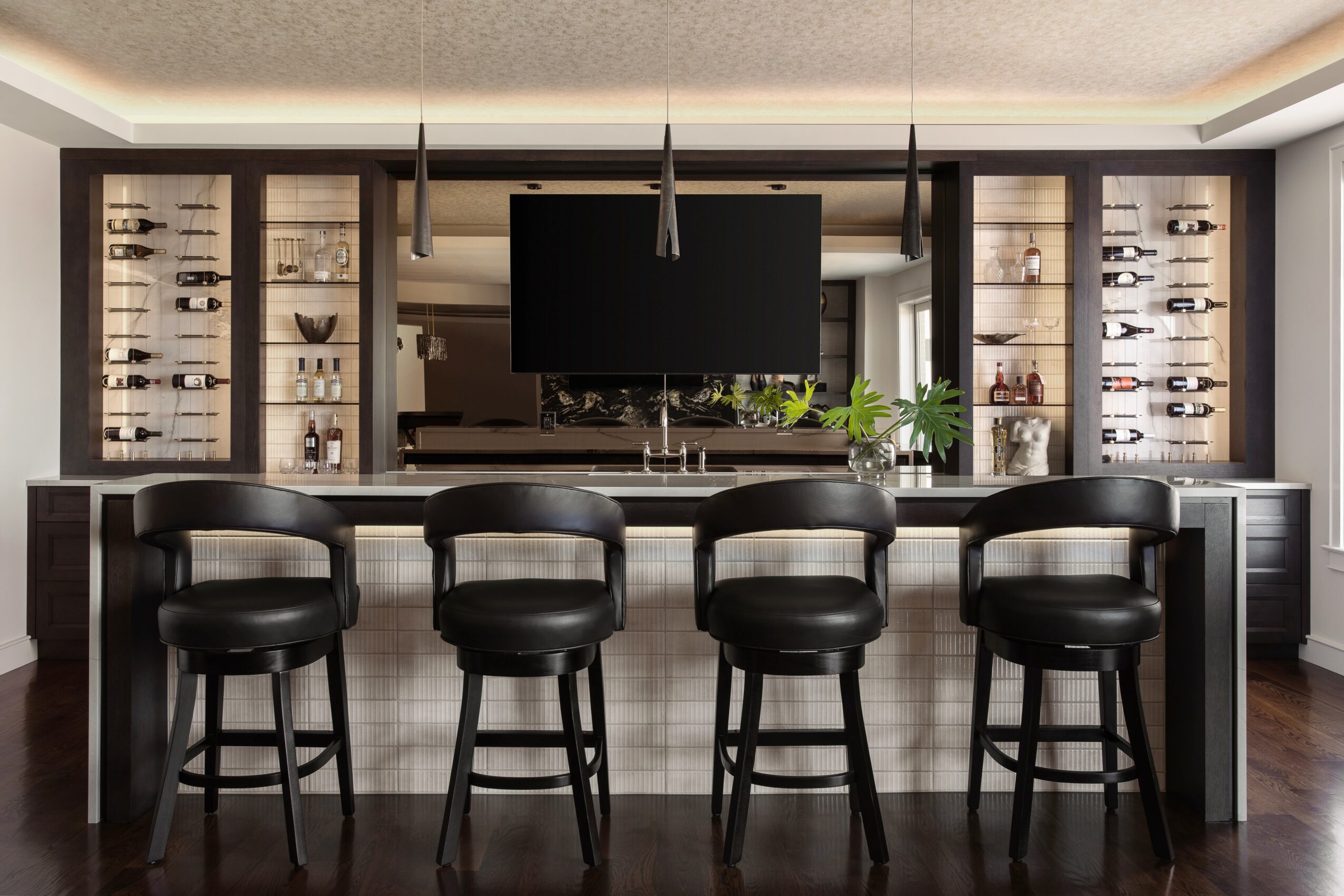 Bar area with four black stools, a large TV, and glass display cabinets with bottles, in a modern room.