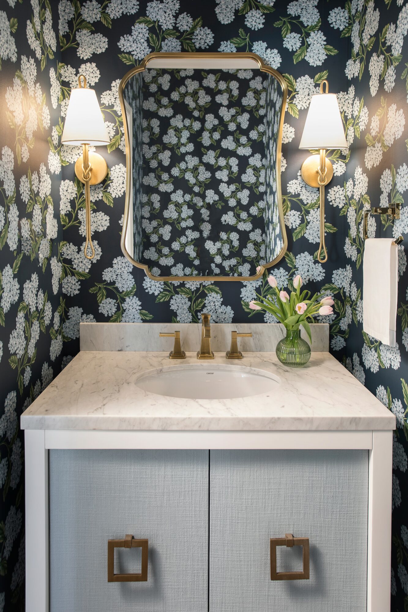 Bathroom vanity with a mirror, floral wallpaper, two wall sconces, and a vase with flowers on the countertop.