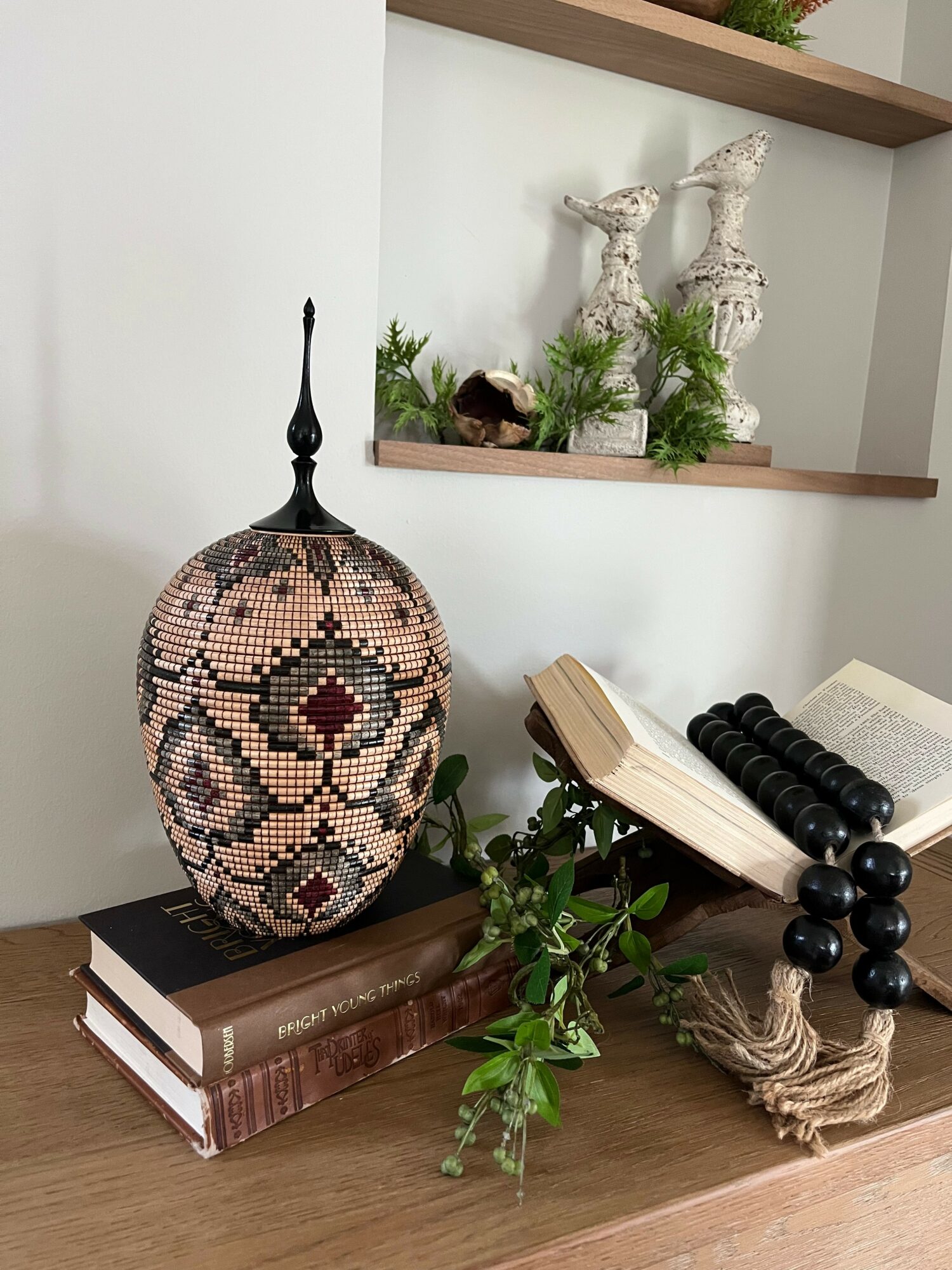 Decorative mosaic vase, open book, black beaded necklace, and green plant on wooden surface with white wall and shelves in background.