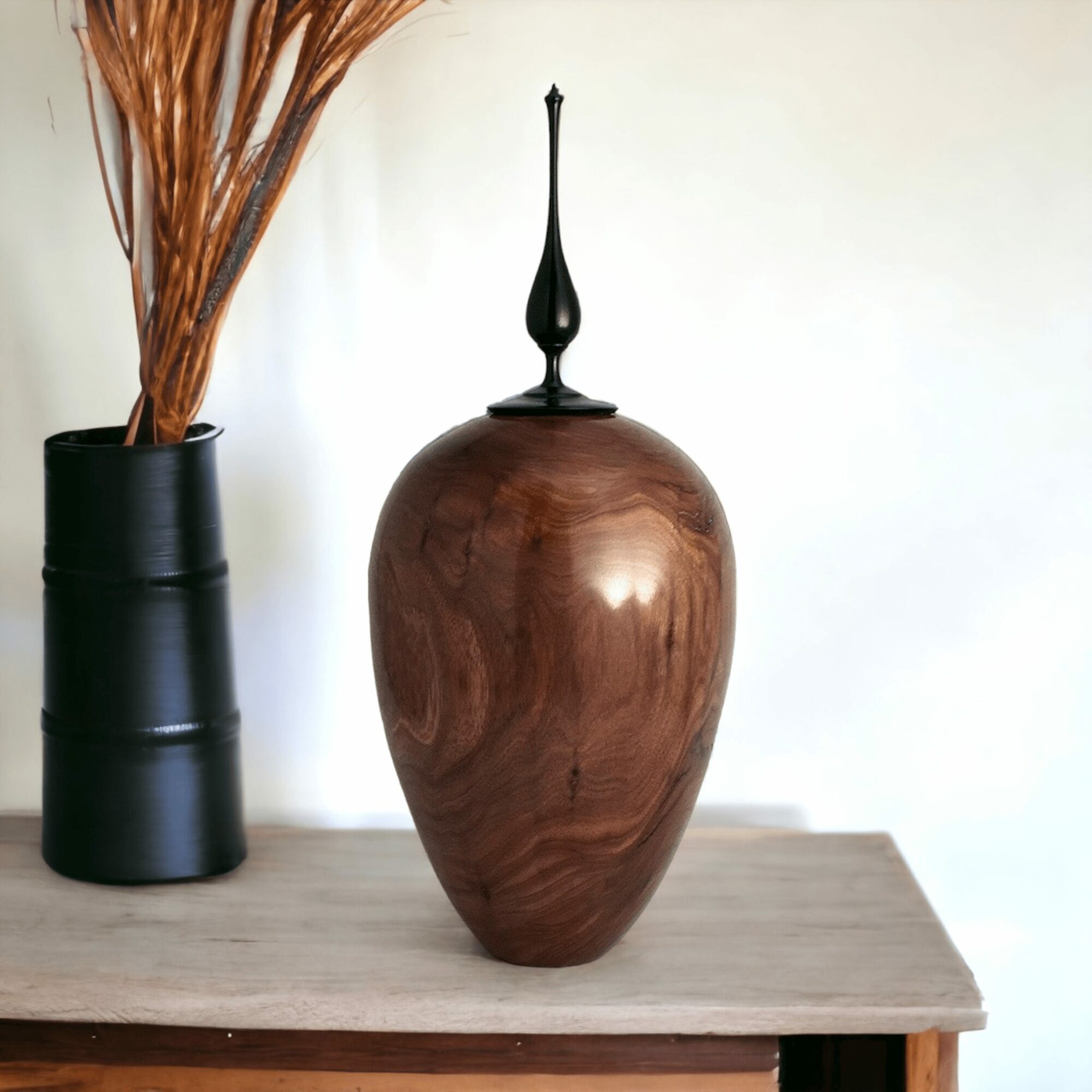Wooden vase with a black decorative finial on top, placed on a wooden surface with a black vase containing dried branches nearby