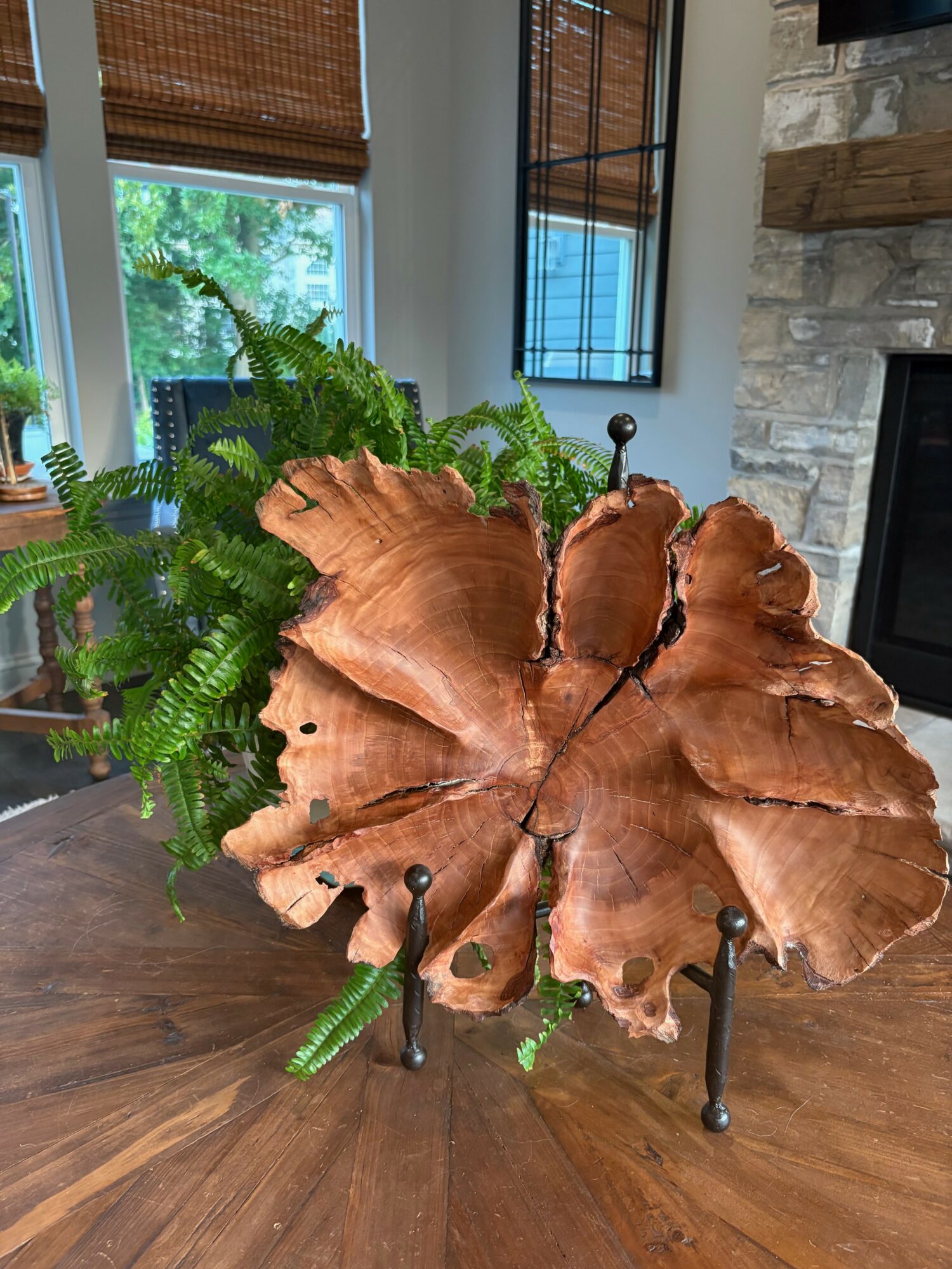 Decorative wooden butterfly sculpture with green fern plant behind it on a wooden table, in a room with windows and stone fireplace.