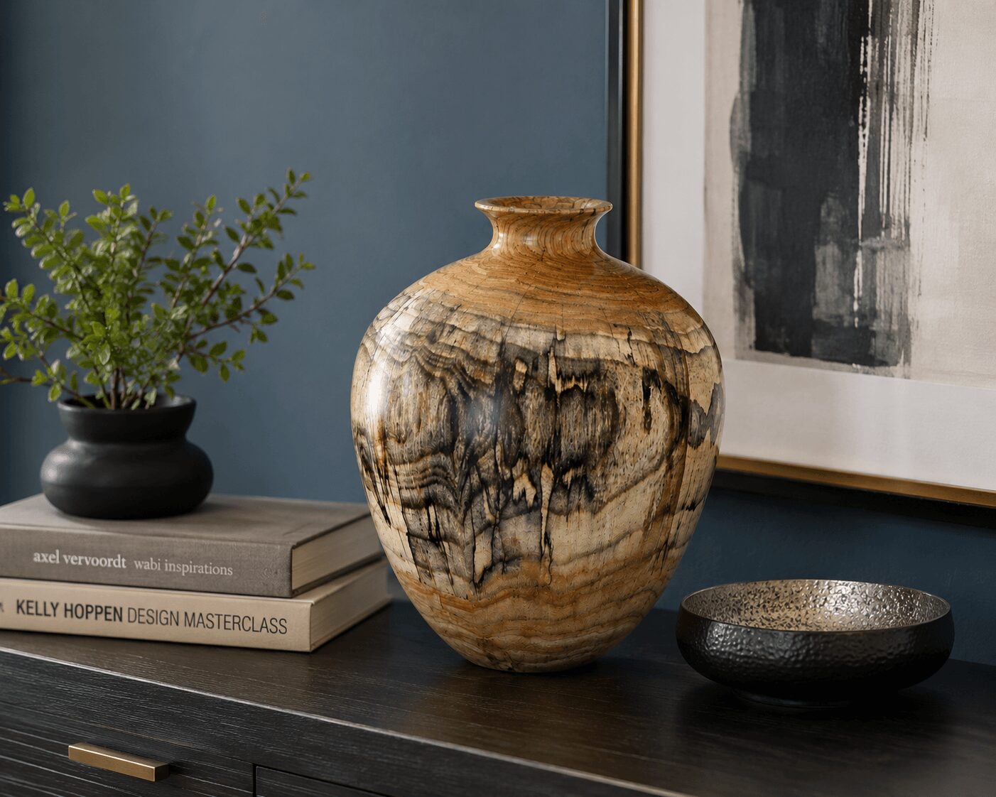Decorative vase with wood grain pattern on a dark surface, next to a black bowl and a potted plant, against a blue wall.