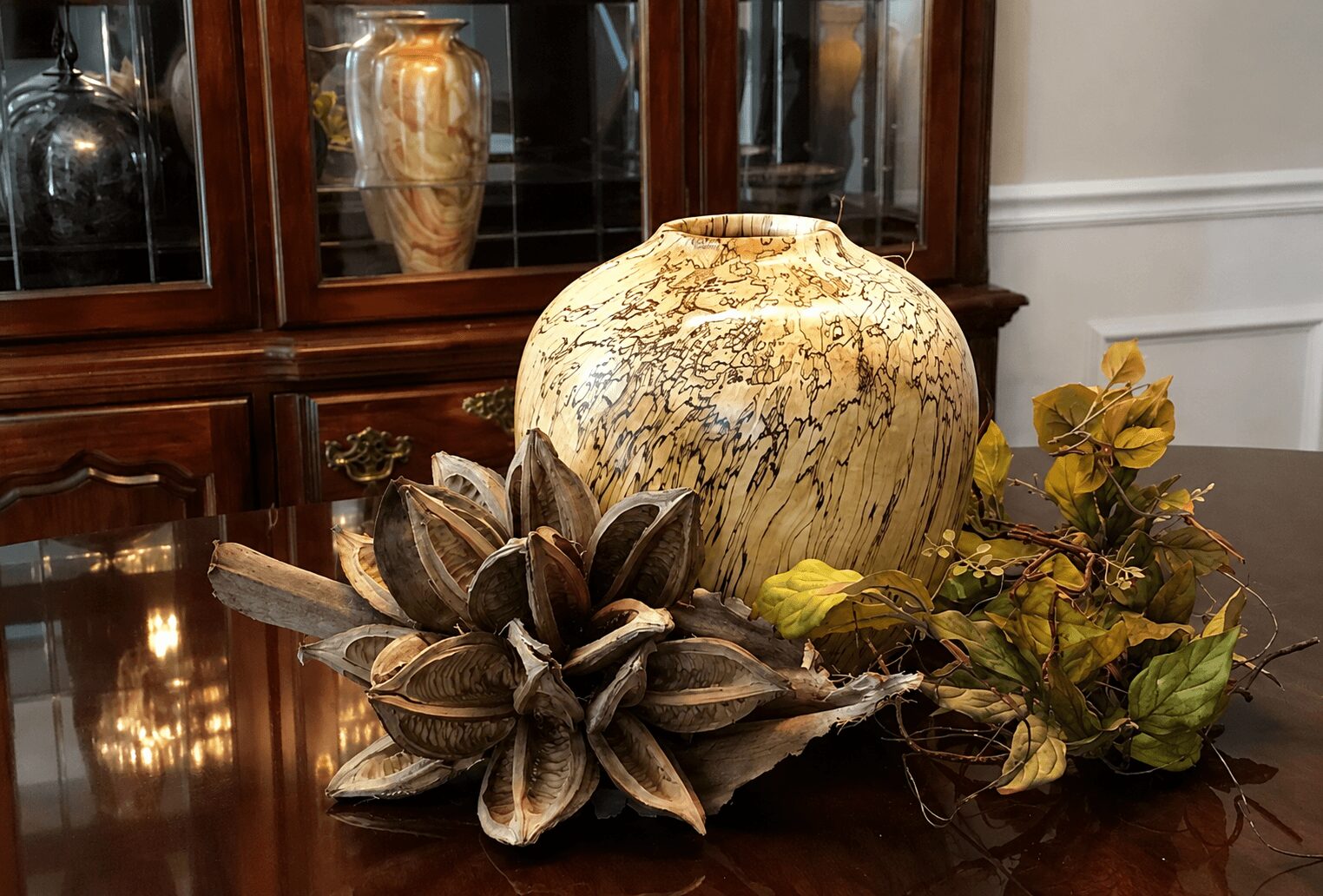 Decorative vase with dried flowers and leaves on a wooden table, with a cabinet in the background.