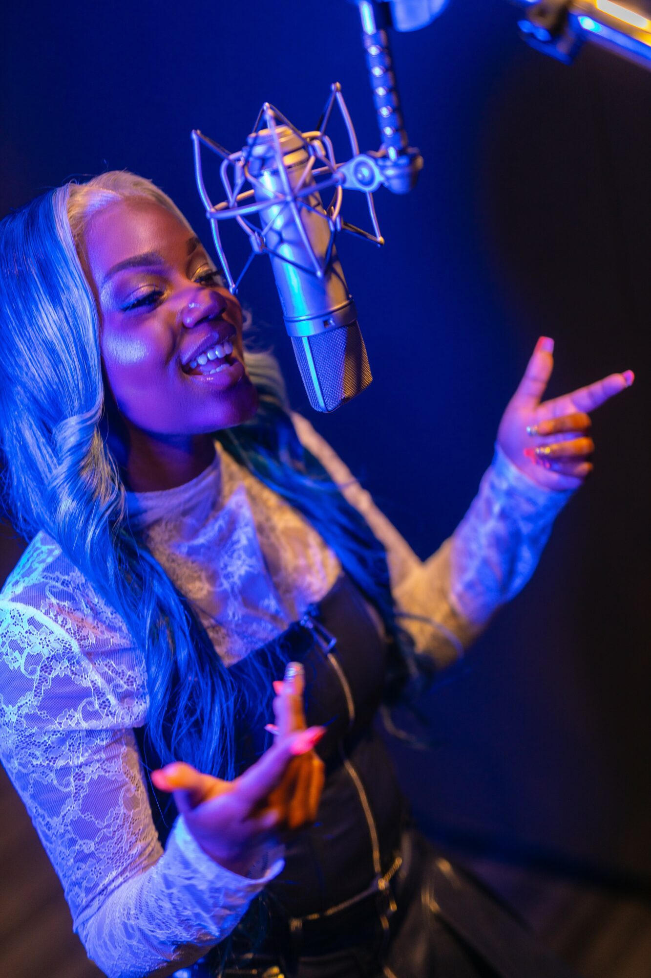 Woman singing into a microphone, smiling, with her hand raised, in a recording studio with blue lighting.
