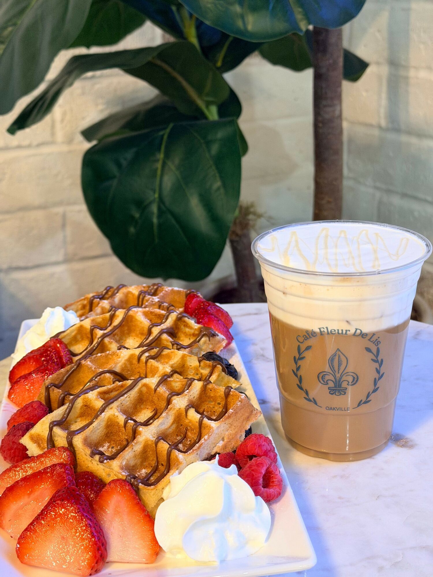 Plate with waffles, strawberries, and whipped cream, next to a coffee drink on a table outdoors.
