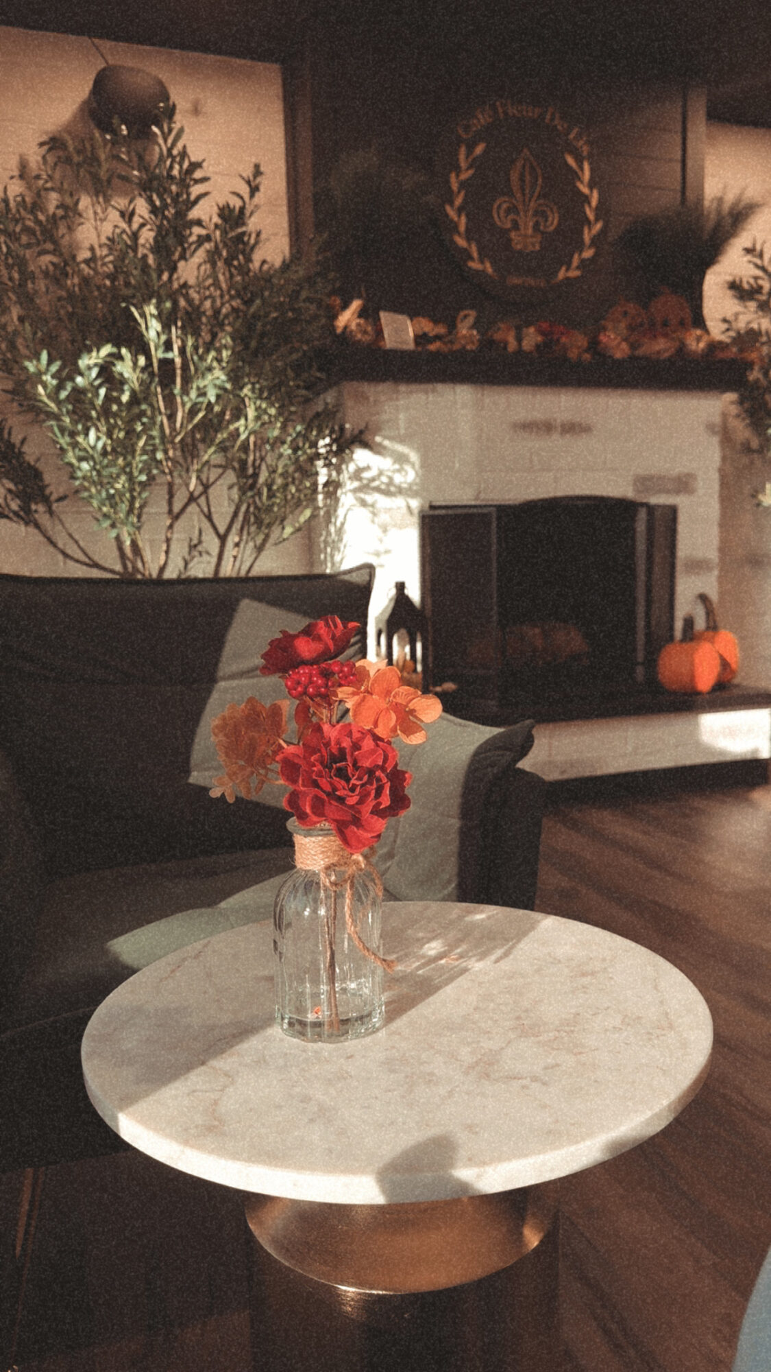 Vase with red flowers on a round table in a cozy room with a fireplace and pumpkins, dim lighting.