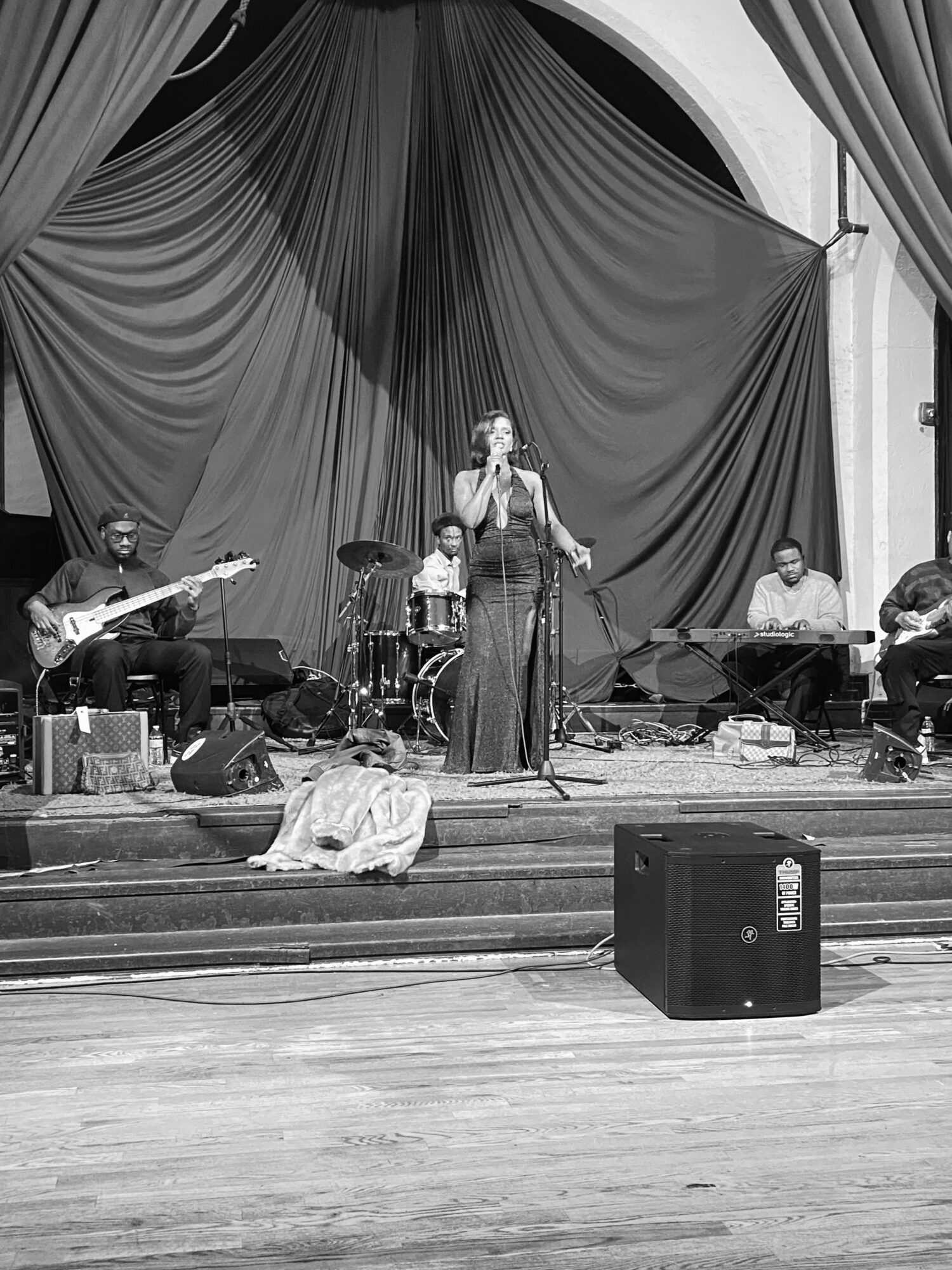 Group of five musicians performing on stage with curtains in the background, one person singing into a microphone, others playing instruments.