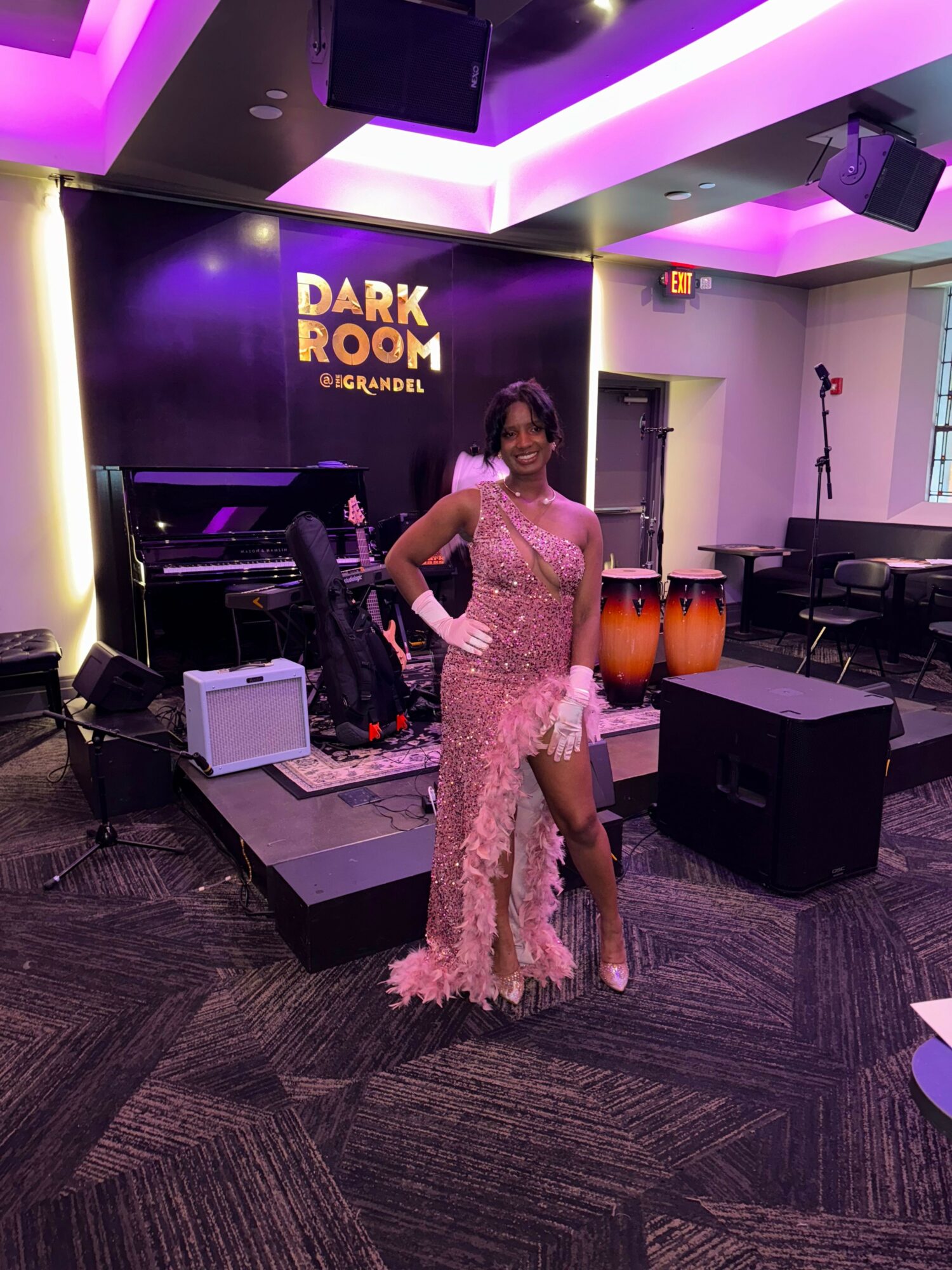 Woman in pink lace dress standing on stage with musical instruments in a dark room with purple lighting.
