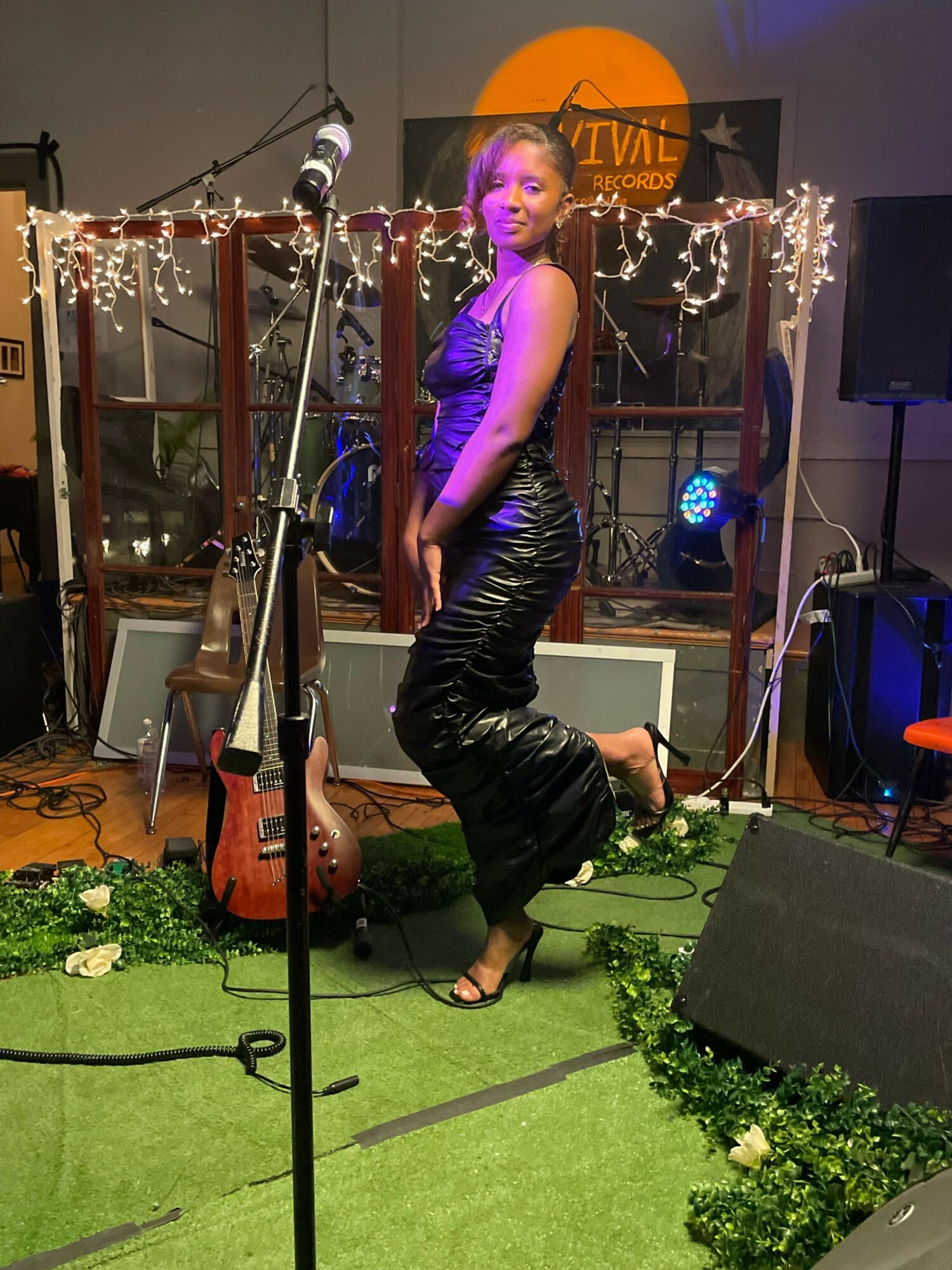 Young woman on stage with microphone, wearing shiny black pants and top, surrounded by musical equipment and string lights.