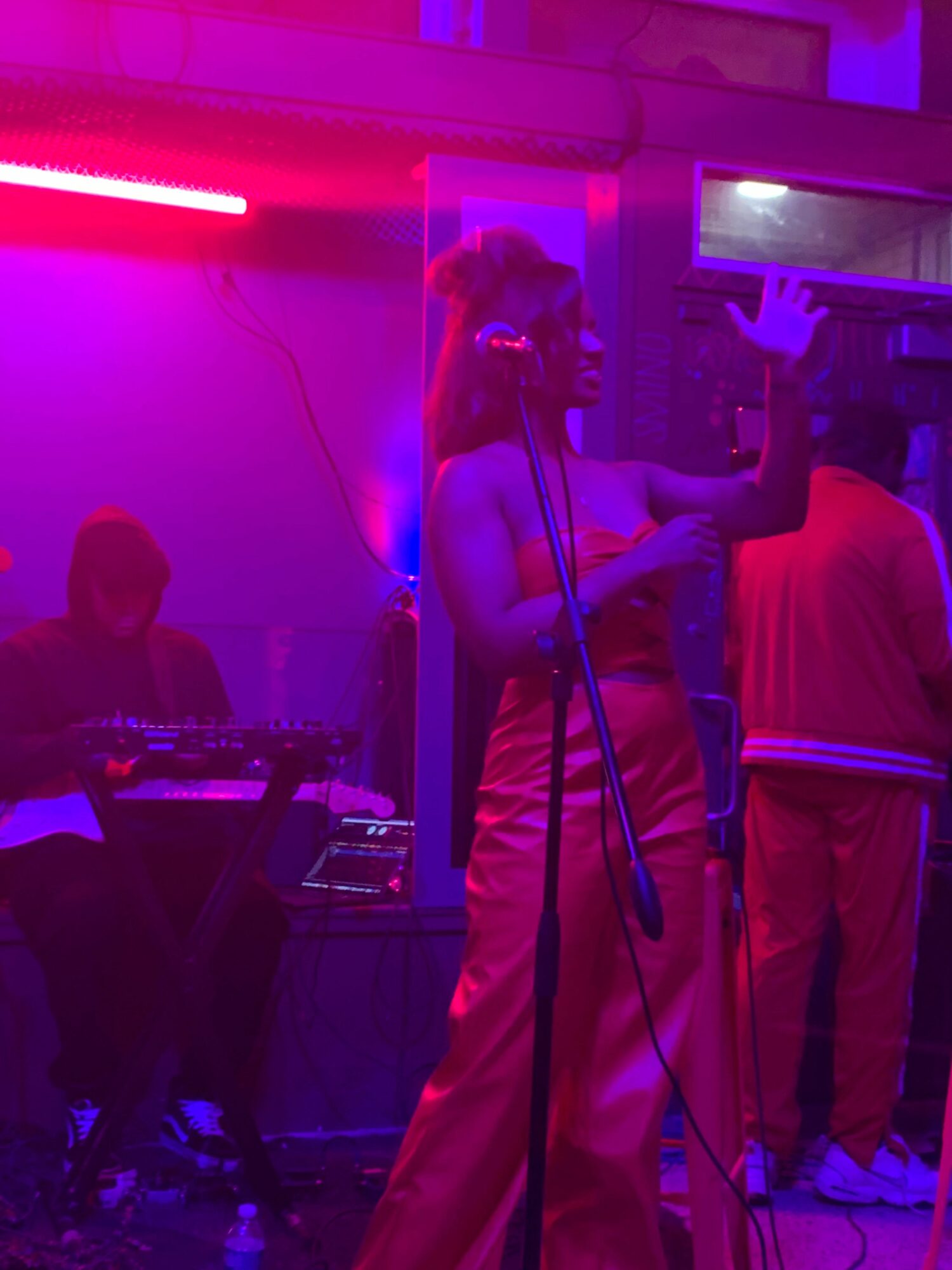 A woman in a pink outfit takes a selfie in a dimly lit room with purple and pink lighting, with a DJ and other people in the background.