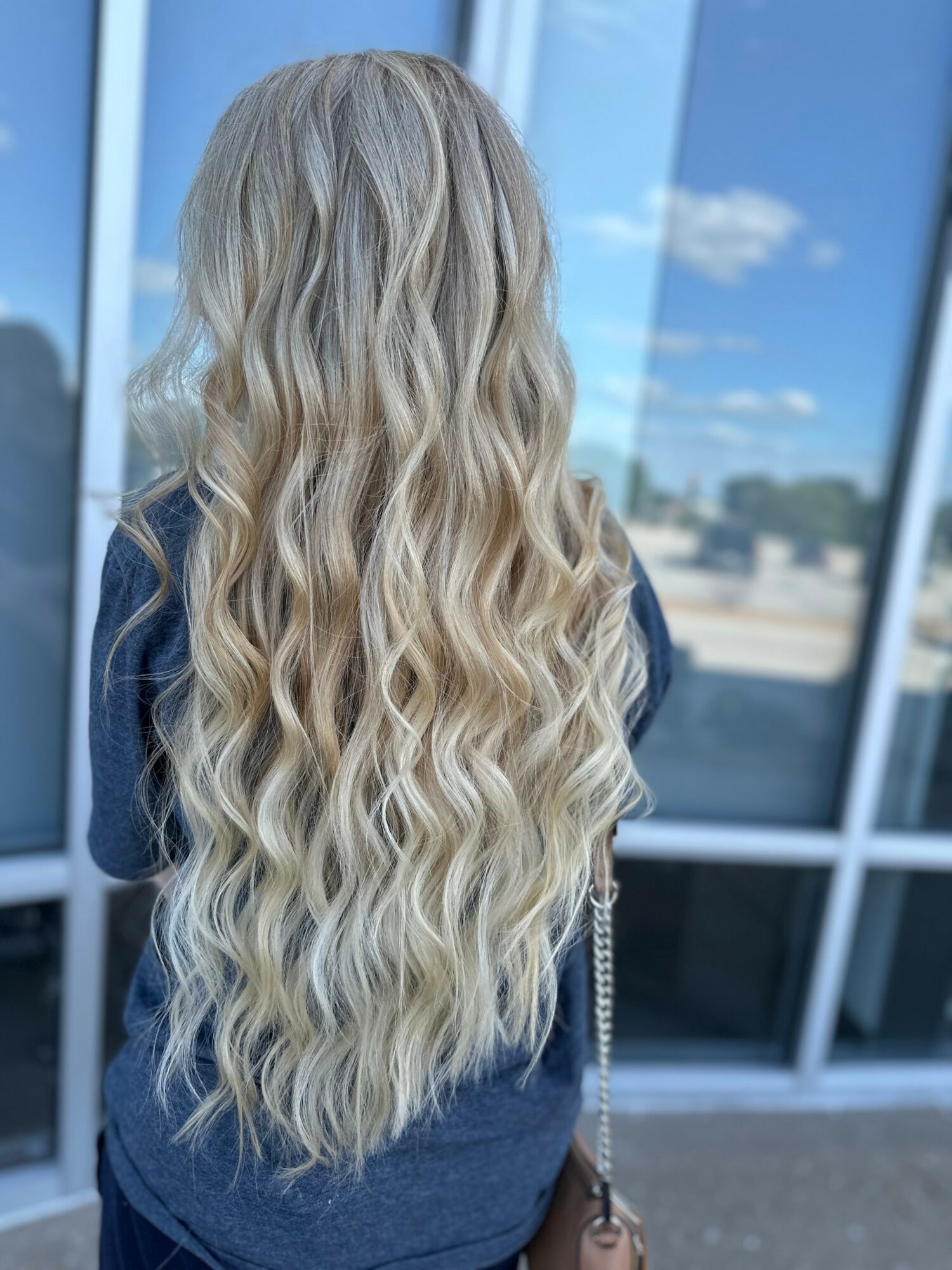 Back view of a person with long, wavy blonde hair standing outside near glass windows, with a blue sky and clouds.