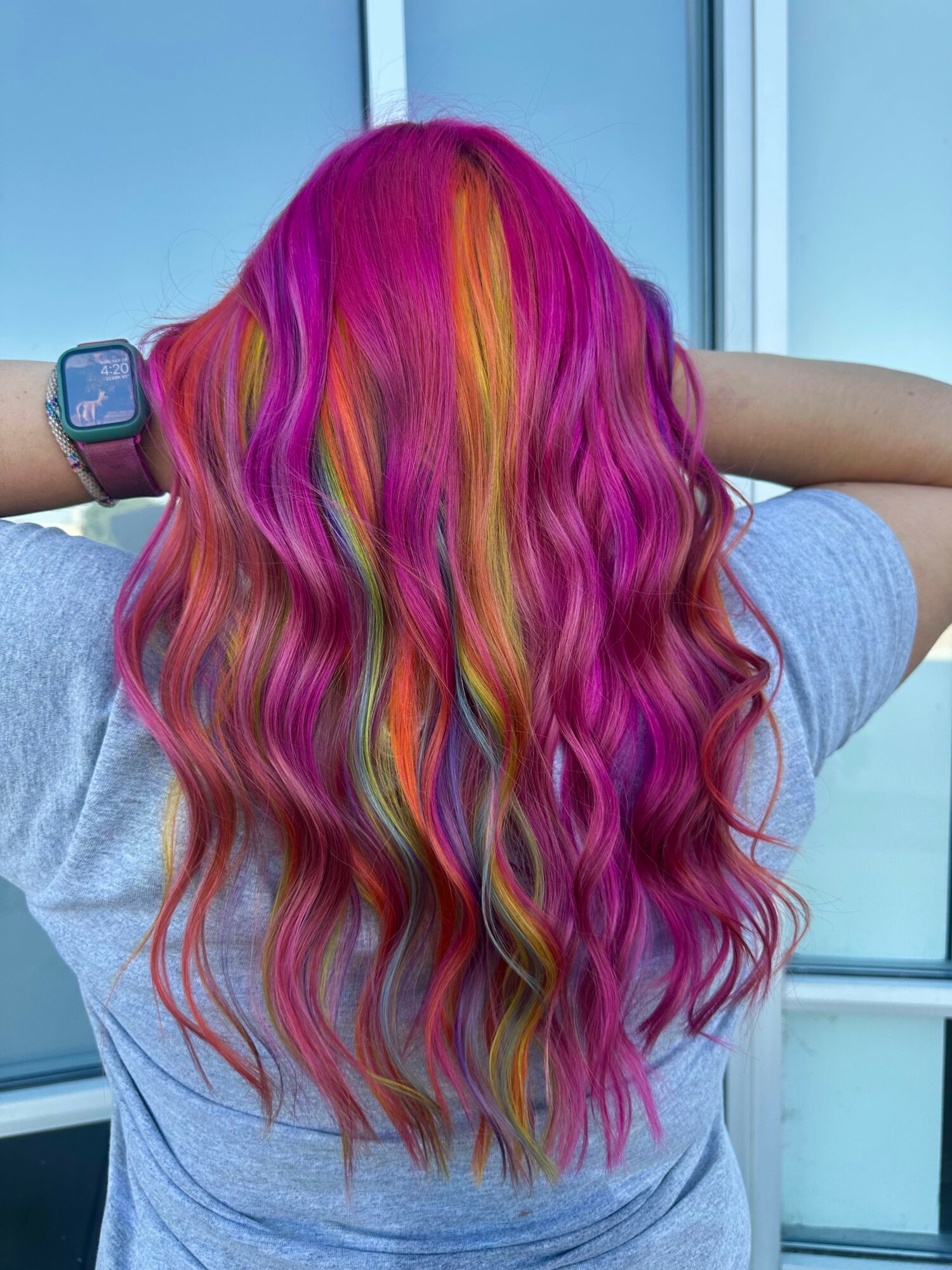 Back view of a person with long, wavy, multicolored hair in pink, orange, yellow, green, and purple hues, wearing a gray shirt.