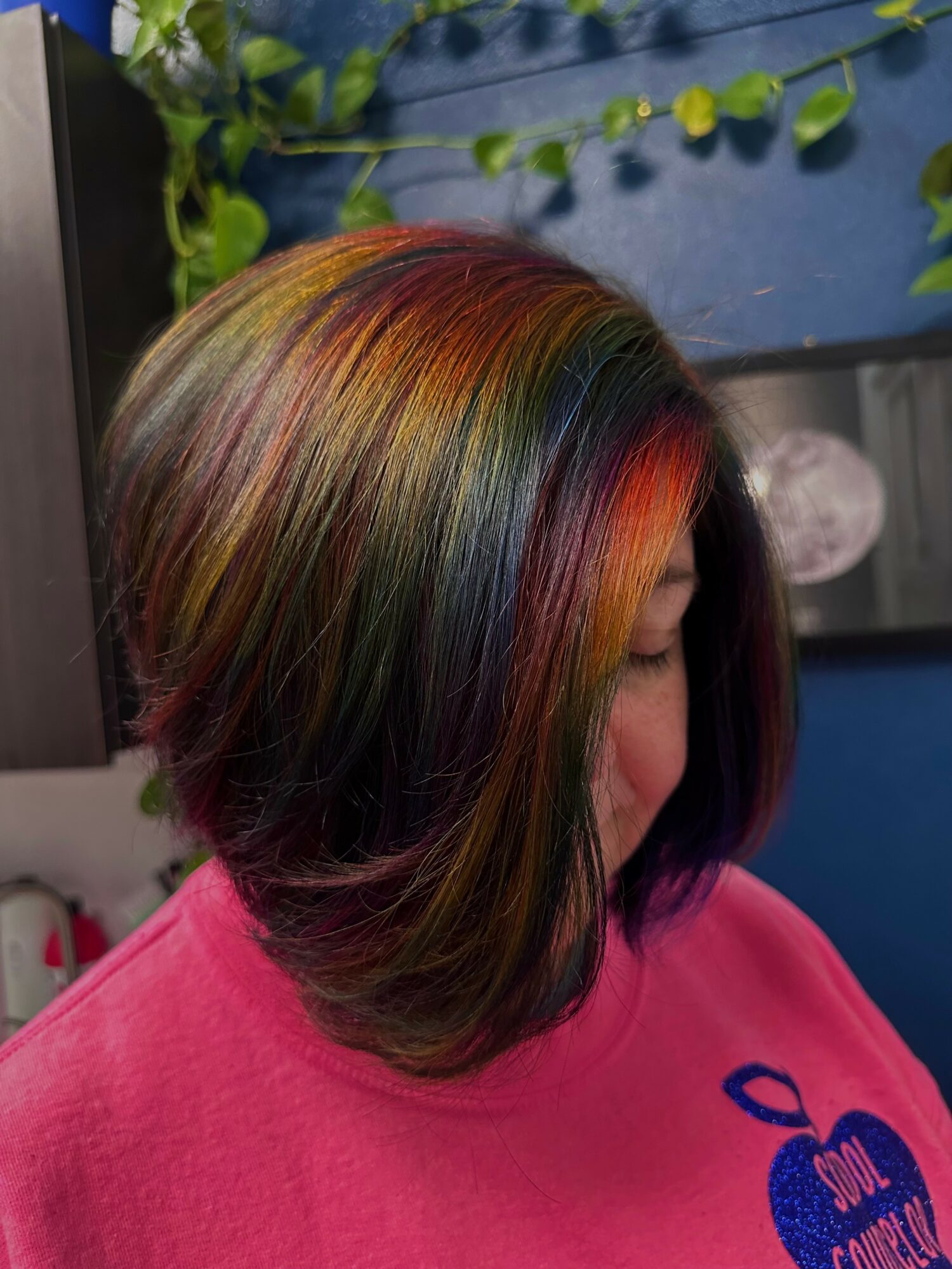 Side view of a woman with a multicolored, layered bob hairstyle, wearing a pink shirt, in a room with blue walls.