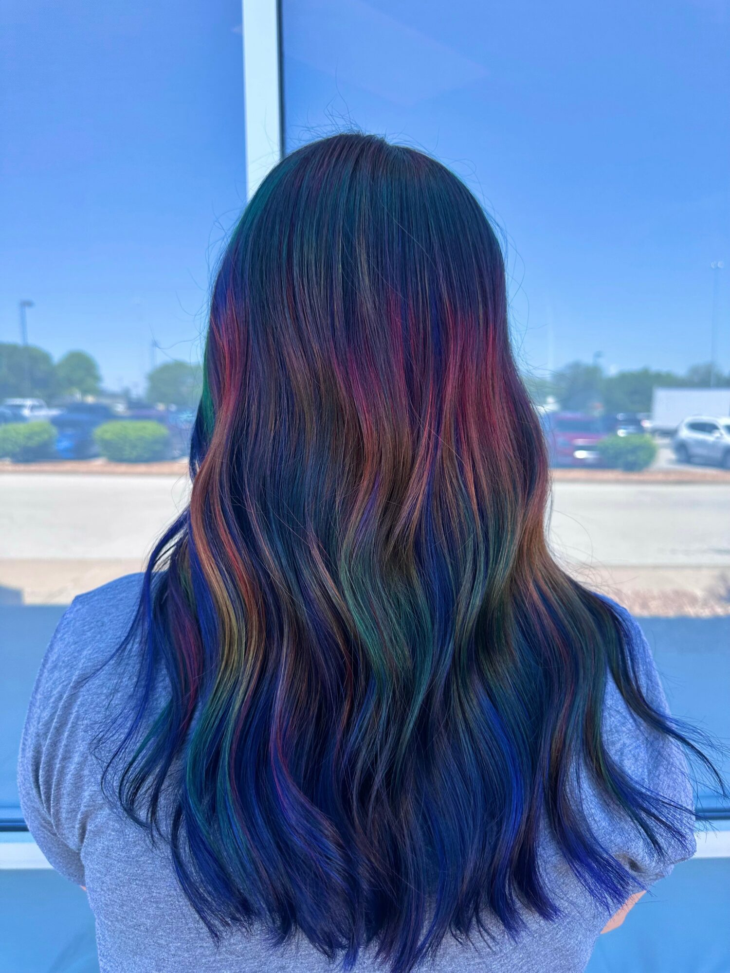 Back view of a person with long, wavy hair dyed in rainbow colors, standing outdoors with a blue sky.