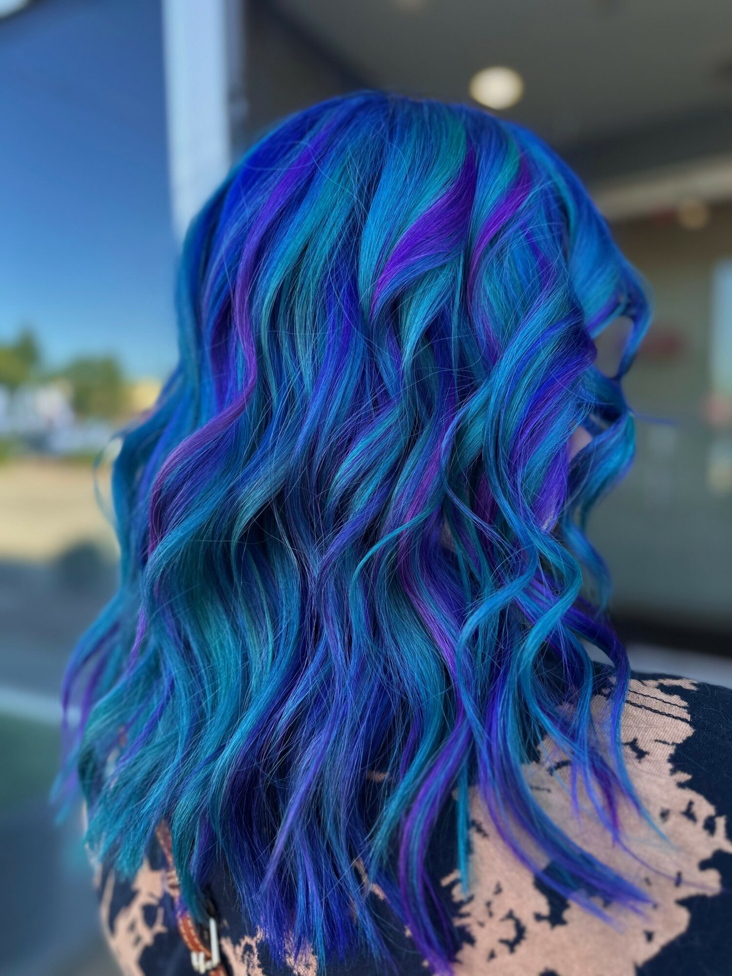 Wavy hair with vibrant blue, purple, and teal colors, styled in loose curls, outdoors with a blurred background.