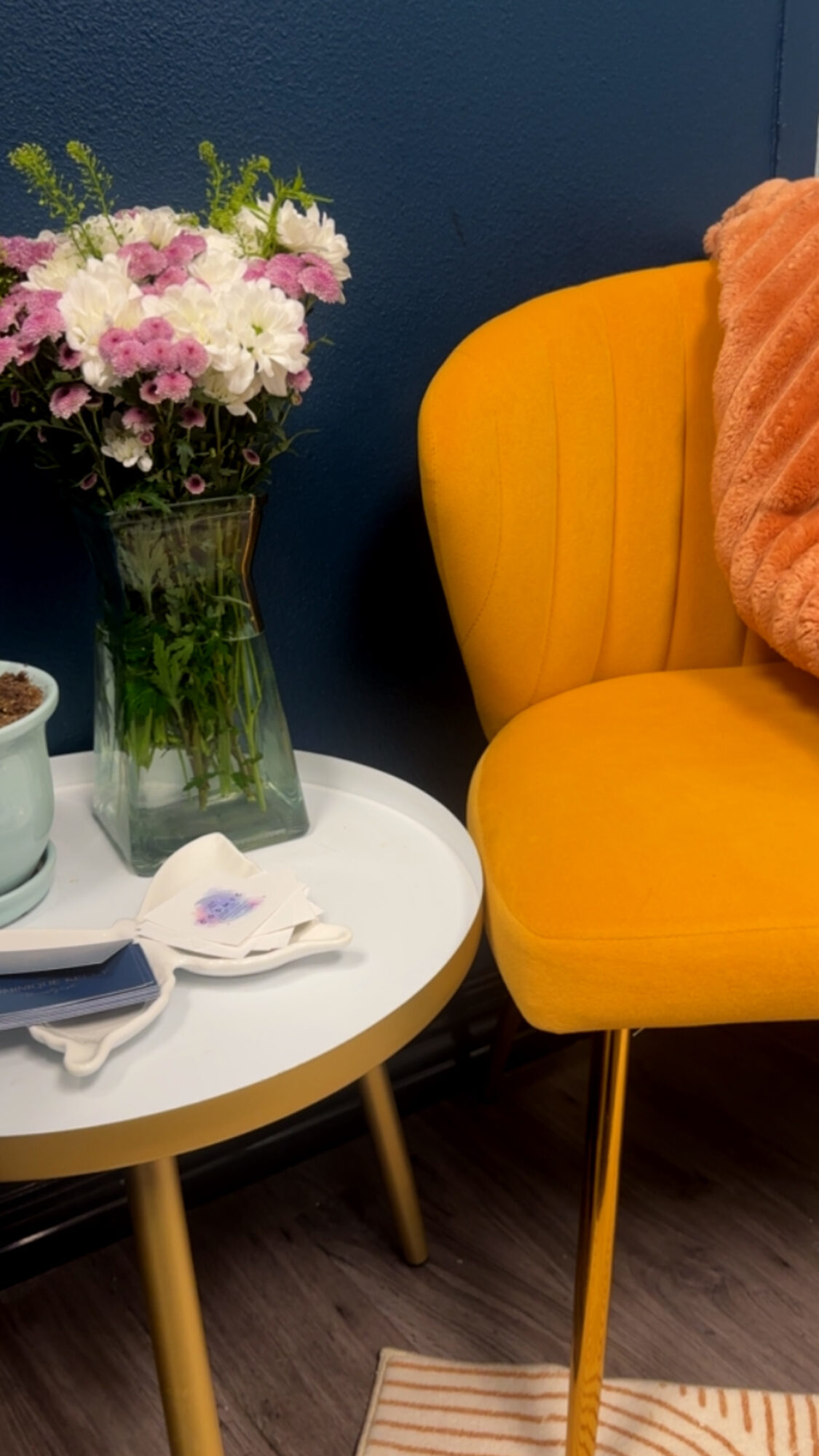 Yellow armchair with orange pillow, white side table with flowers and a cup, dark blue wall background.