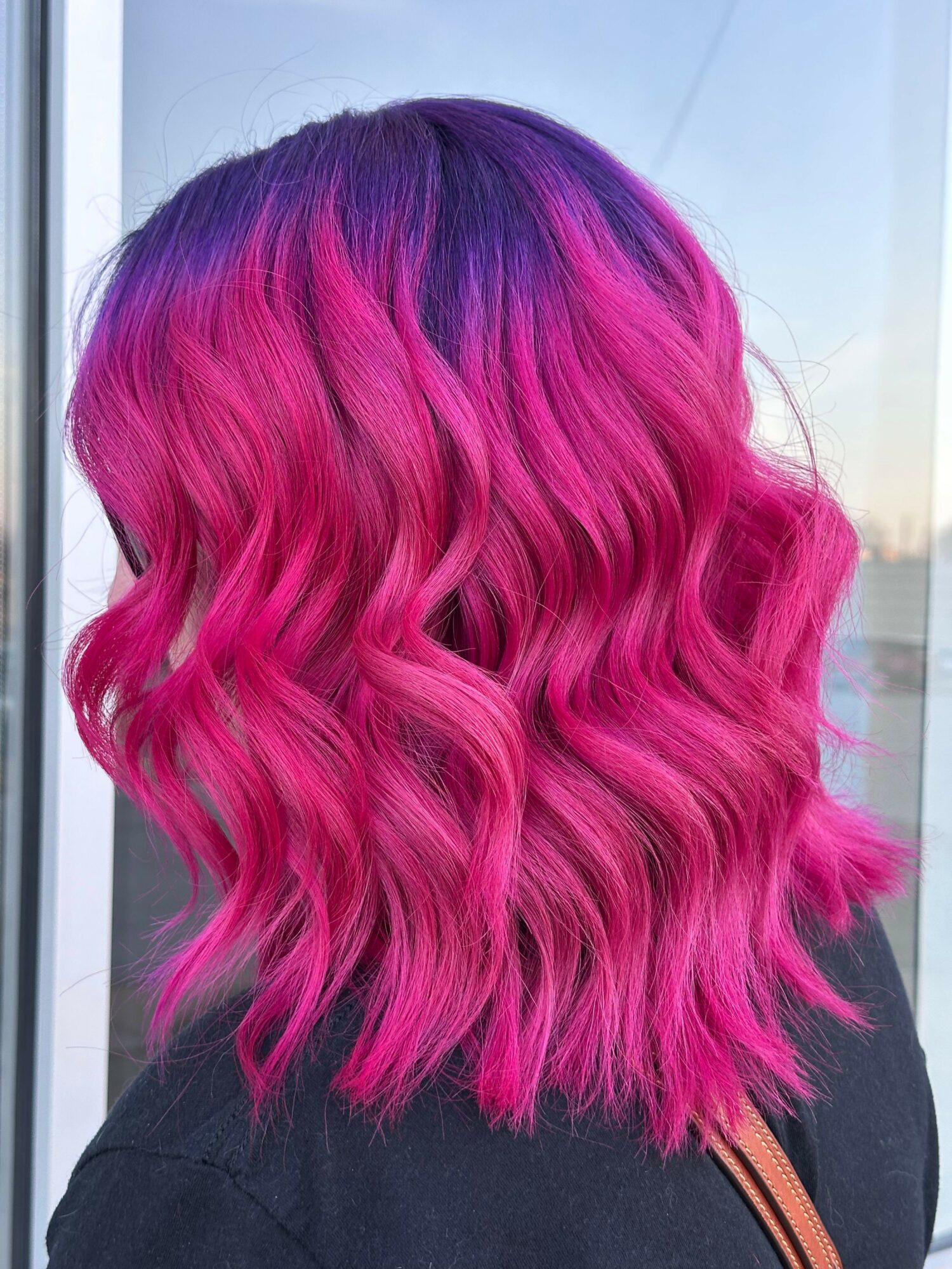 Person with vibrant pink and purple wavy hair, viewed from the side, against a light background.