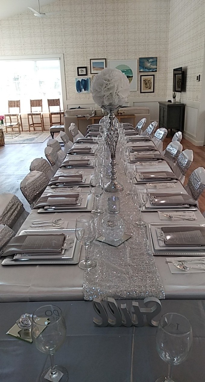 Long dining table set with plates, glasses, and napkins, decorated with a tall floral centerpiece, in a bright room.