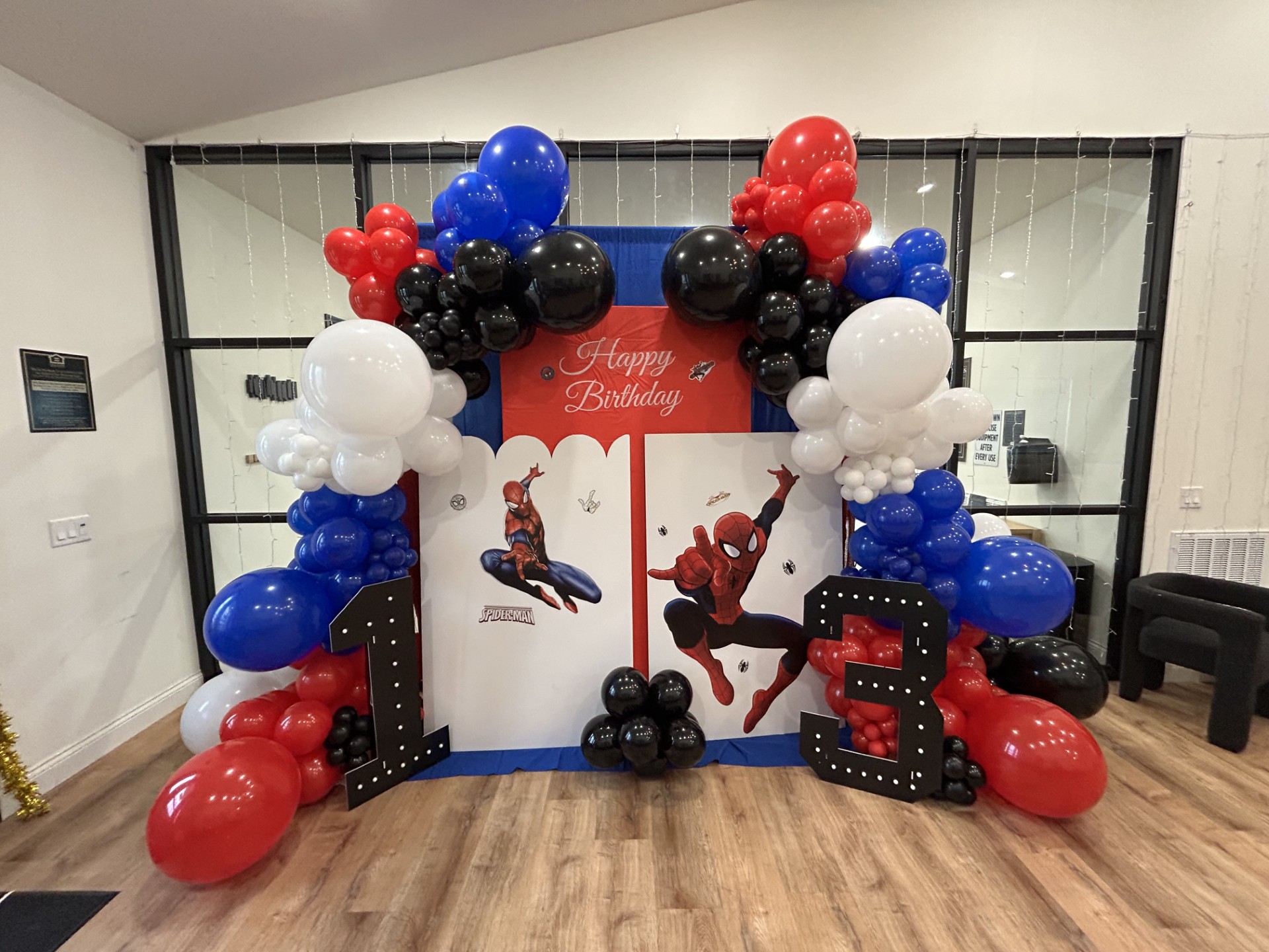 Colorful balloon arch with red, blue, black, and white balloons surrounds a birthday backdrop with Spider-Man theme, celebrating a third birthday.