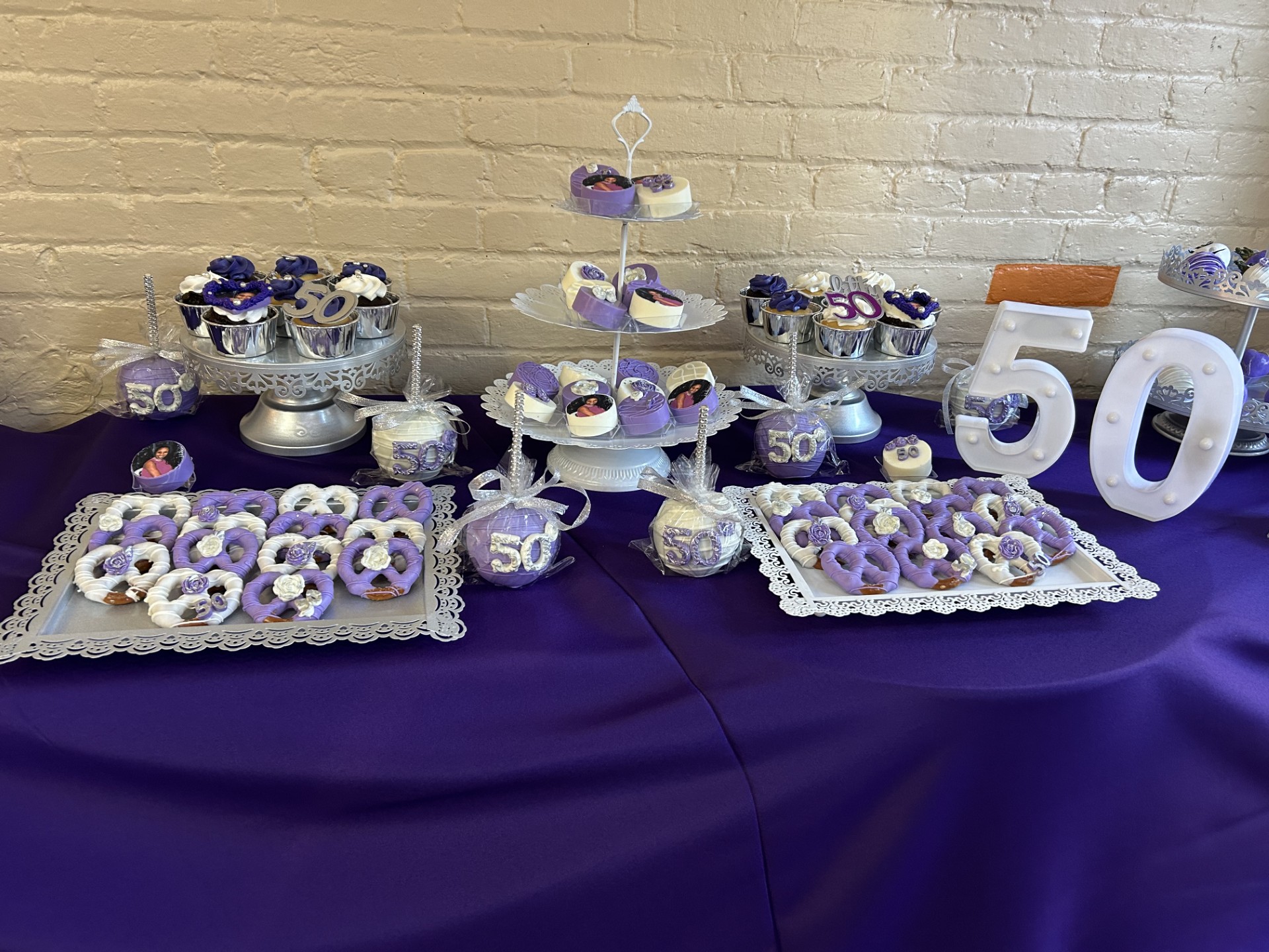 Table with purple and white cupcakes, cookies, and decorations celebrating a 50th birthday, against a brick wall background.