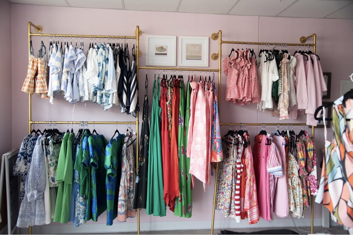 Clothing racks with various colorful dresses and shirts in a boutique, pink walls, framed pictures, and a mirror visible.