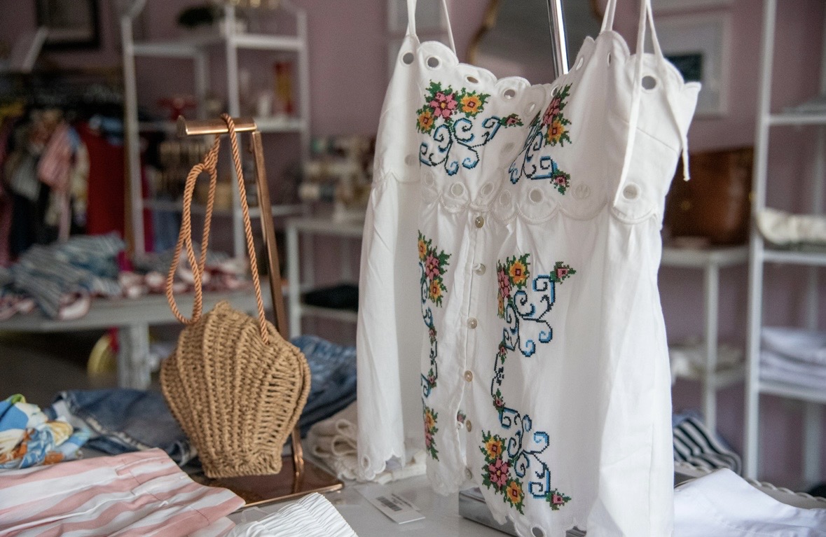 White bags with colorful floral embroidery hanging in a store, with clothing and woven bag in background.