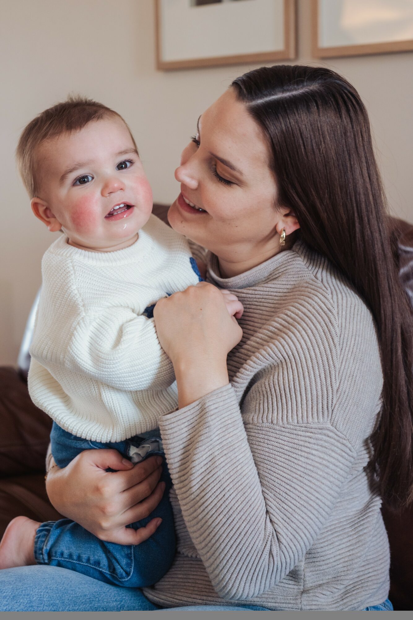 Woman holding smiling child indoors, both looking at each other, woman wearing a gray sweater, child in white sweater.